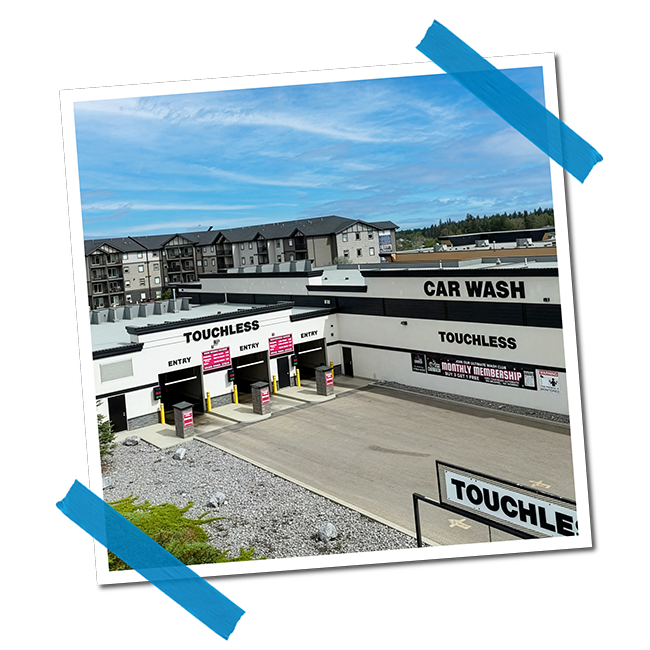 Exterior view of a touchless car wash facility with multiple bays, signage for entry and monthly membership, surrounded by a parking lot and some greenery, under a cloudy sky.