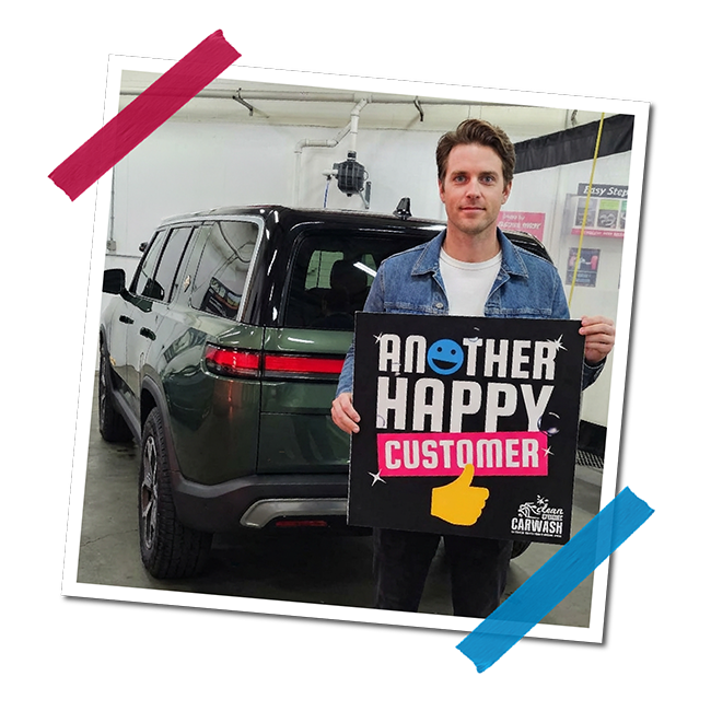 A man holding a sign that says 'Another Happy Customer' with a smiley face and a thumbs-up icon in front of a black and green vehicle at a car wash, with a white wall and equipment in the background.