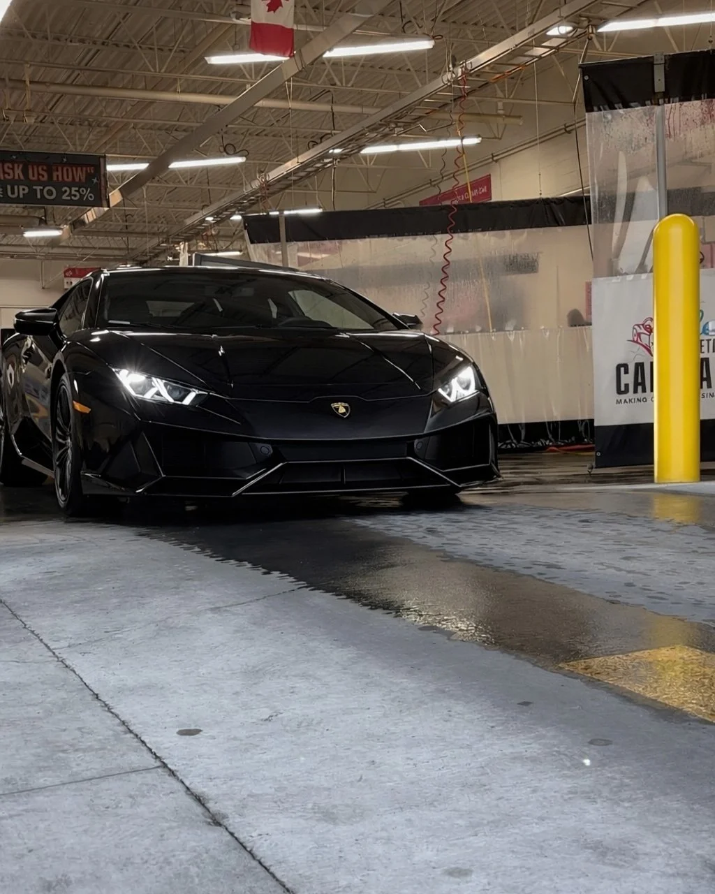 Black Lamborghini sports car inside an auto wash bay with water on the ground and yellow protective post on the right.
