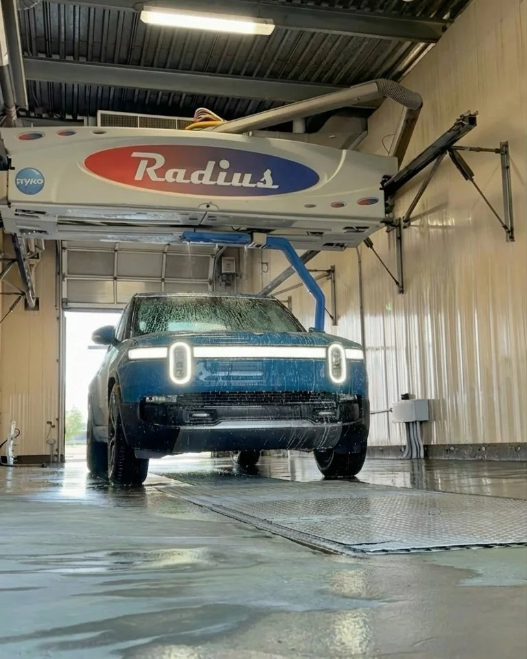 An electric vehicle being washed in a car wash bay with a large Radius branded water cleaning system overhead.