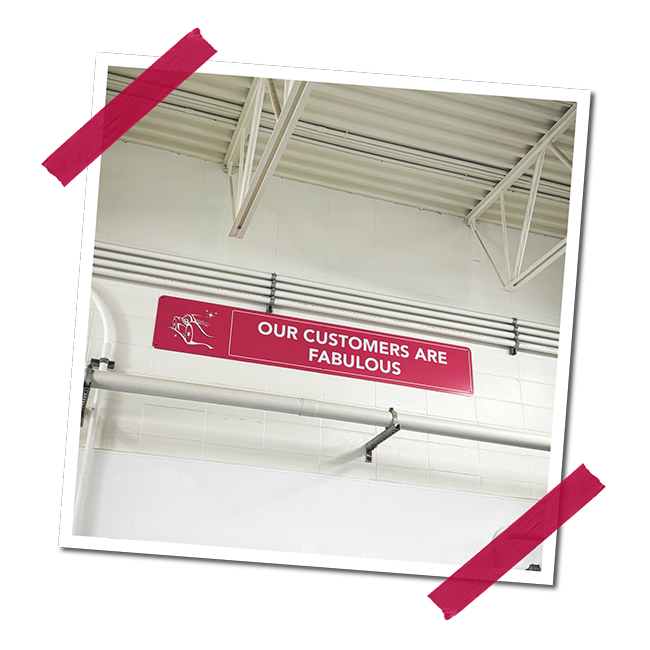 Red sign that reads 'Our customers are fabulous' hanging on a white wall in a warehouse or industrial space.