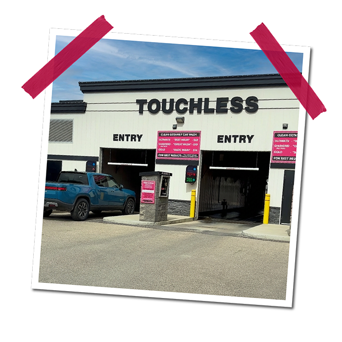 The exterior of a Touchless car wash facility with two entry lanes, a blue truck parked beside it, and signs displaying car wash services.