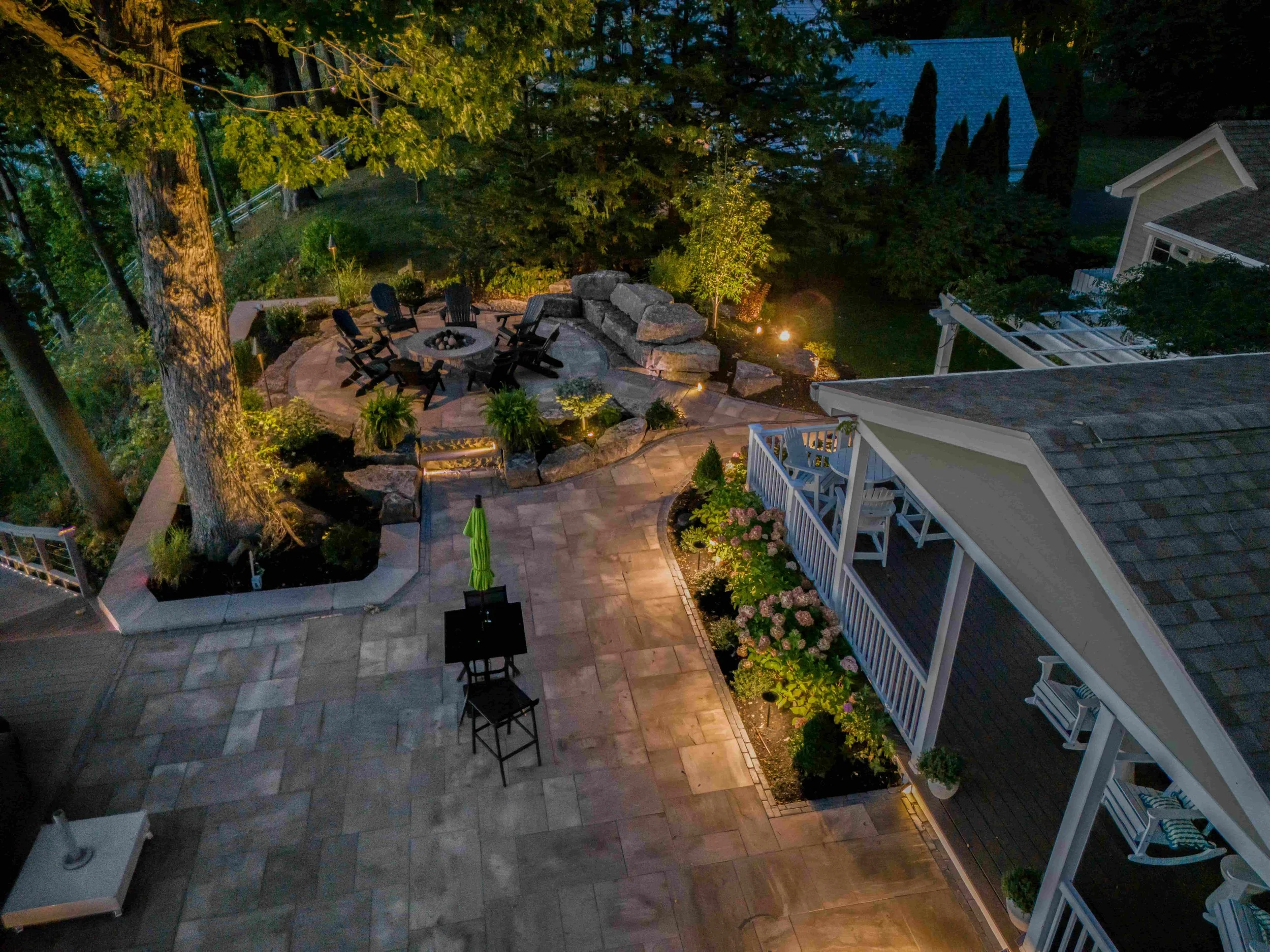 Aerial view of a backyard patio at night with outdoor seating around a fire pit, landscaping, trees, and a house with a porch.