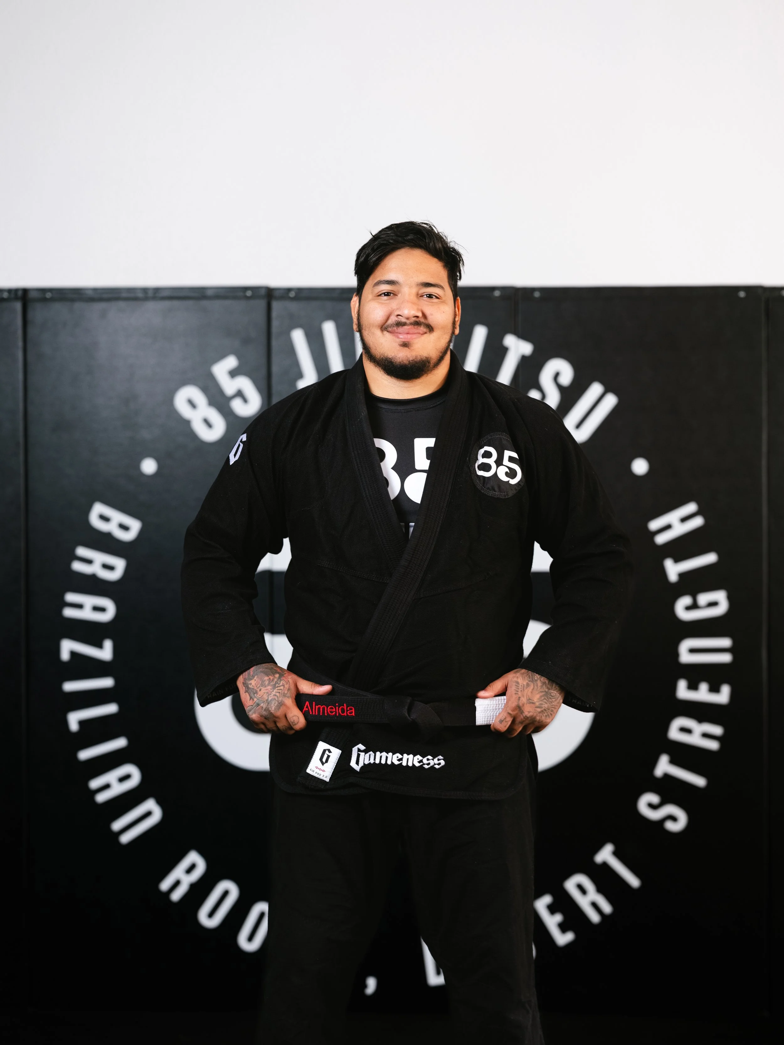 A young man with short dark hair, a beard, and tattoos on his arms, smiling and wearing a black Brazilian Jiu-Jitsu gi standing in front of a wall with a circular logo and text.
