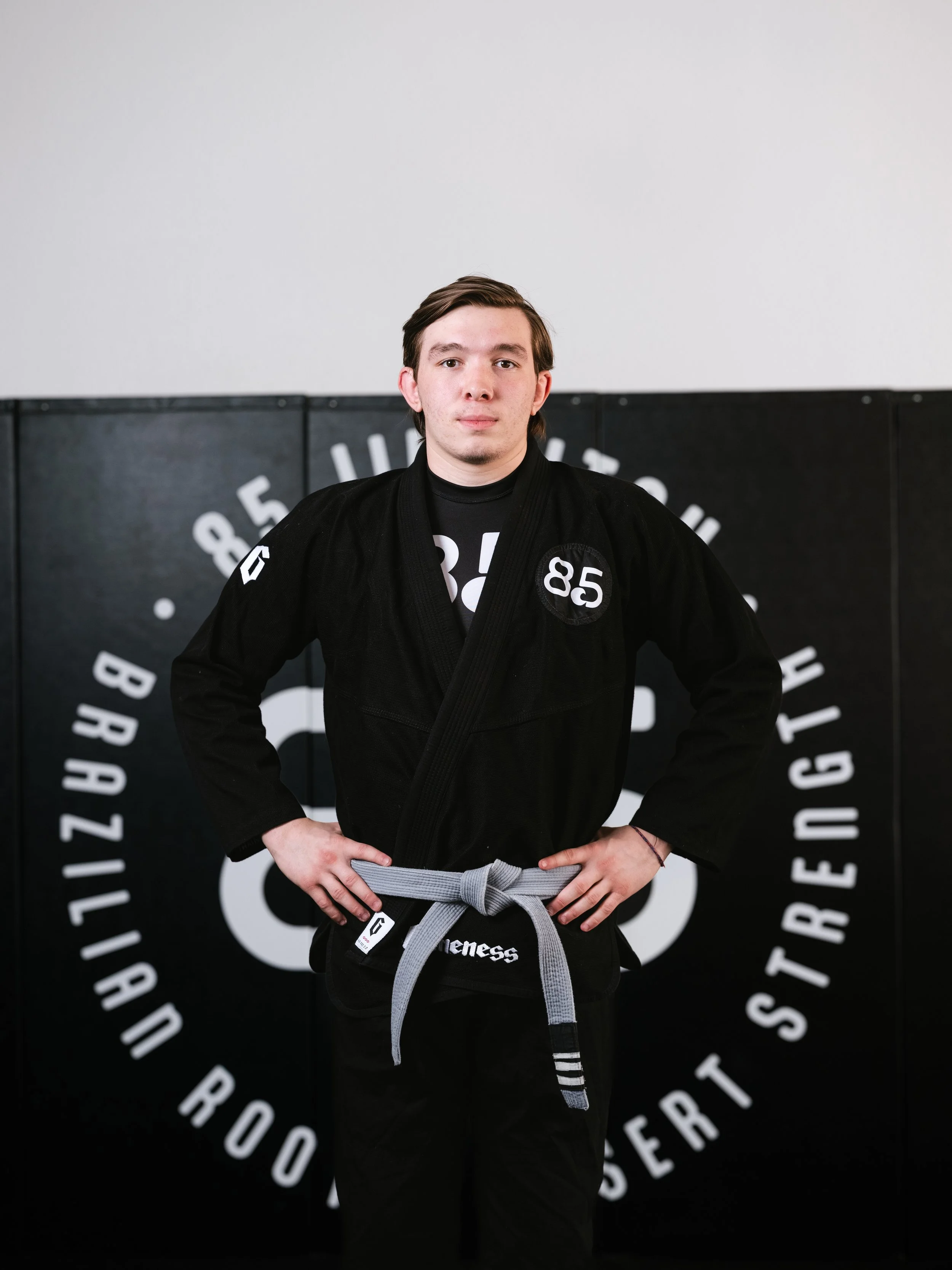 A young man in a judo gi with a gray belt stands in front of a circular logo on the wall. The logo contains the words 'Black Belt' and 'World' among other partially visible words, all in white text on a black background.