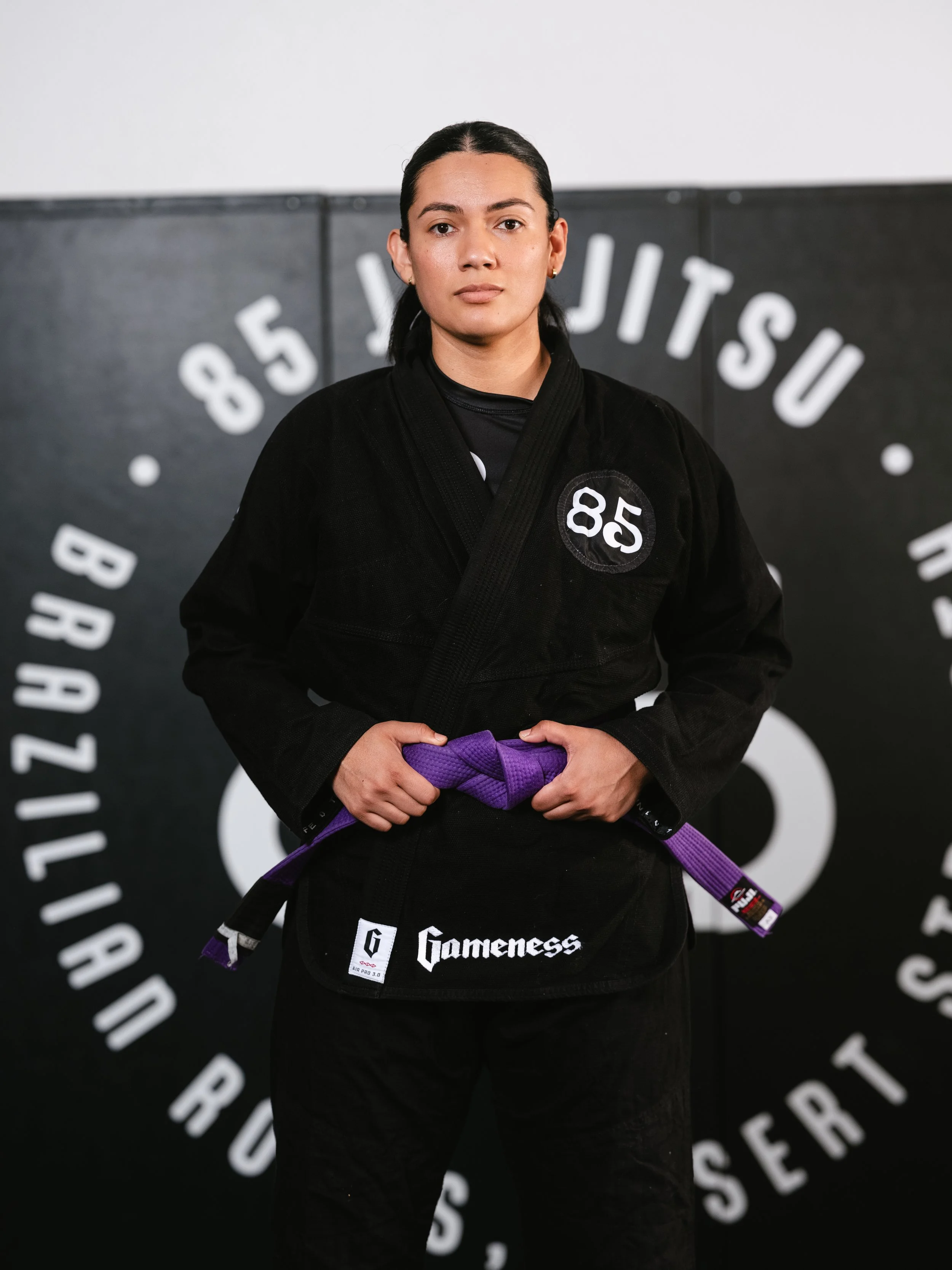 Black and white photo of a woman with dark hair tied back, wearing a black martial arts uniform with a logo on the chest, holding a martial arts belt, standing in front of a background with the words 'Gameness' and other partially visible text.