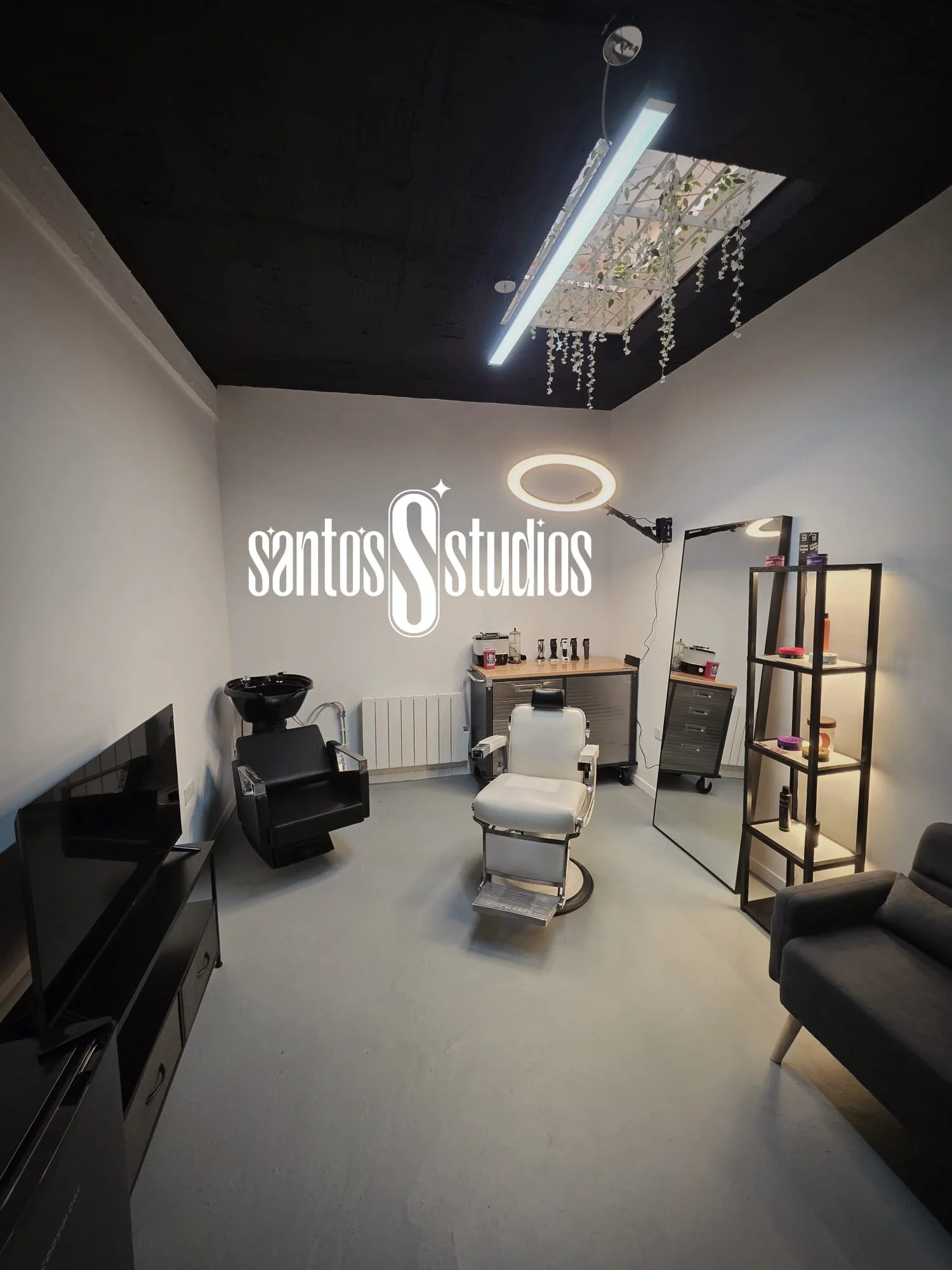 A modern hair salon or barber shop with a white styling chair, black wash basin, mirror, shelving unit, and a ceiling with a skylight and hanging plants. The salon has minimalist decor with a television, black armchair, and various hair products. The logo 'santos Studios' is prominently displayed on the wall.