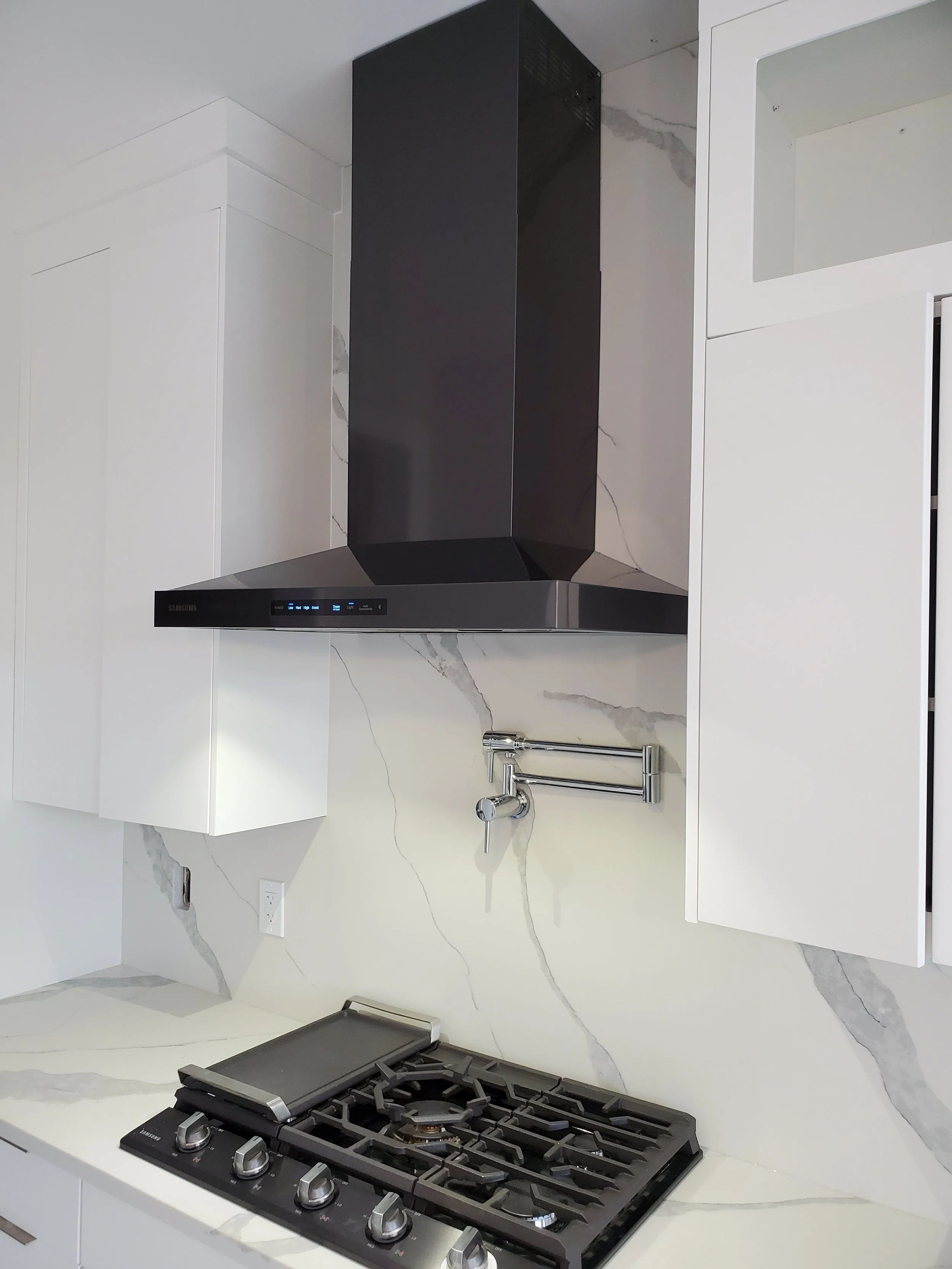 Modern kitchen with white cabinets, marble backsplash, black and stainless range hood, gas stovetop, and wall-mounted pot filler faucet. range exhaust