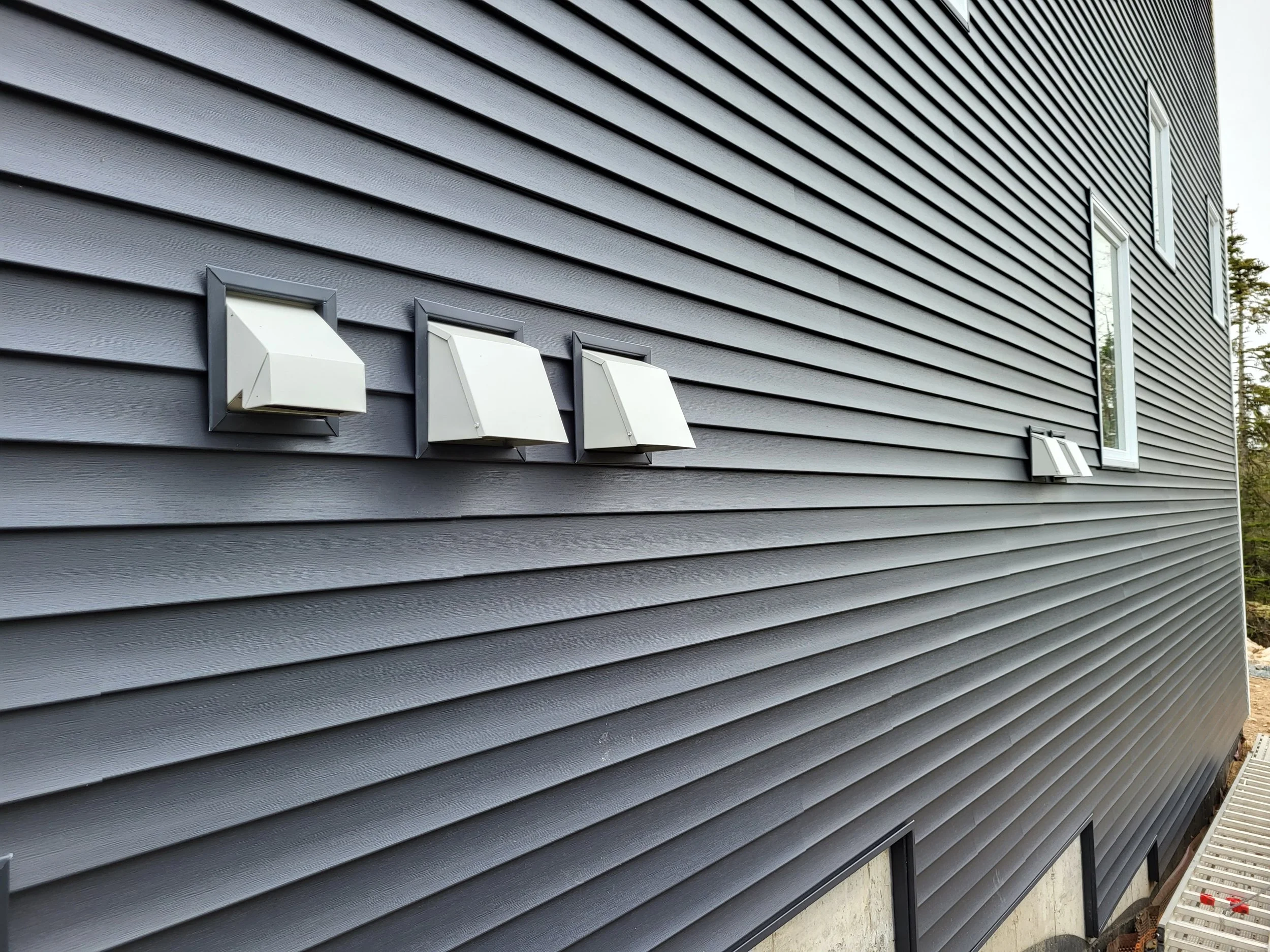 The side of a house with dark gray siding, several white exterior vents, and two windows. effecient air flow and exhaust