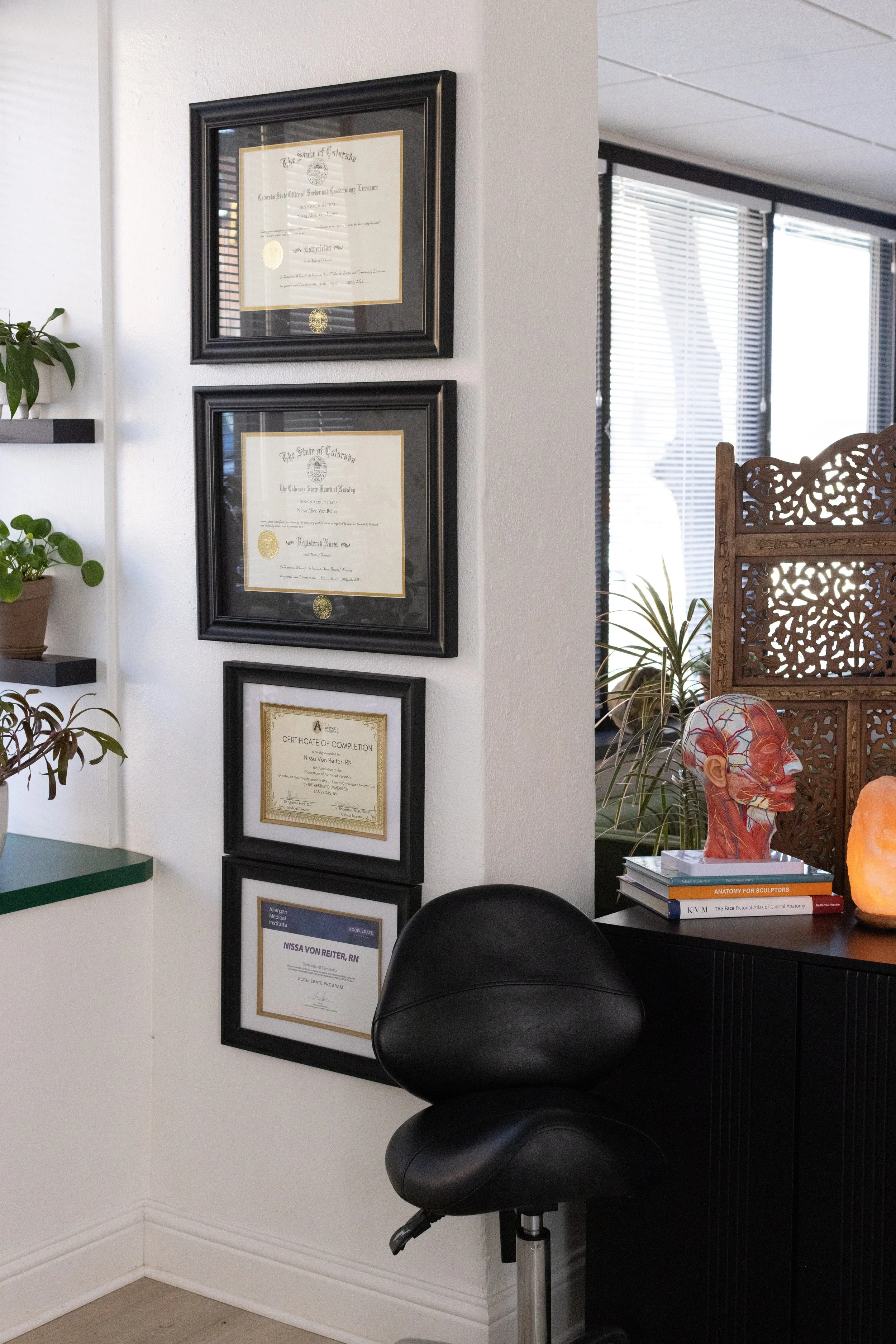 Wall with four framed certificates and diplomas, black swivel chair in front, plants on shelves to the side, decorative wood partition, anatomical sculpture and salt lamp on desk.