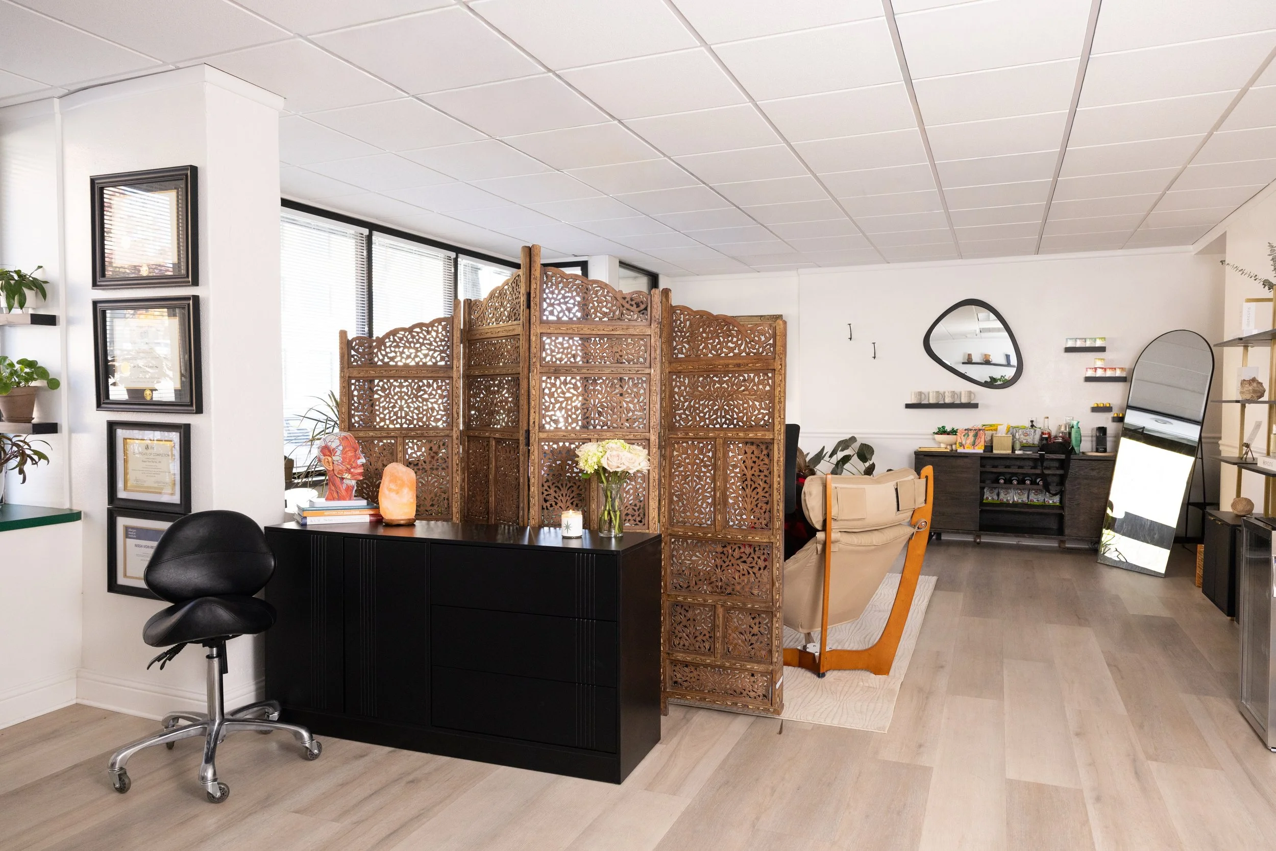 Interior of a modern living space featuring a black desk with a lamp and a flower vase, decorative wooden room divider, beige armchair, large mirror, shelves with plants and artwork, and a sideboard with decor and drinks.
