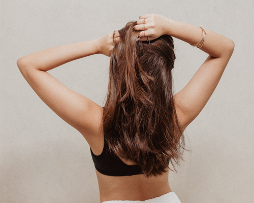 Back view of a woman with long brown hair, wearing a black top, holding her hair with both hands.
