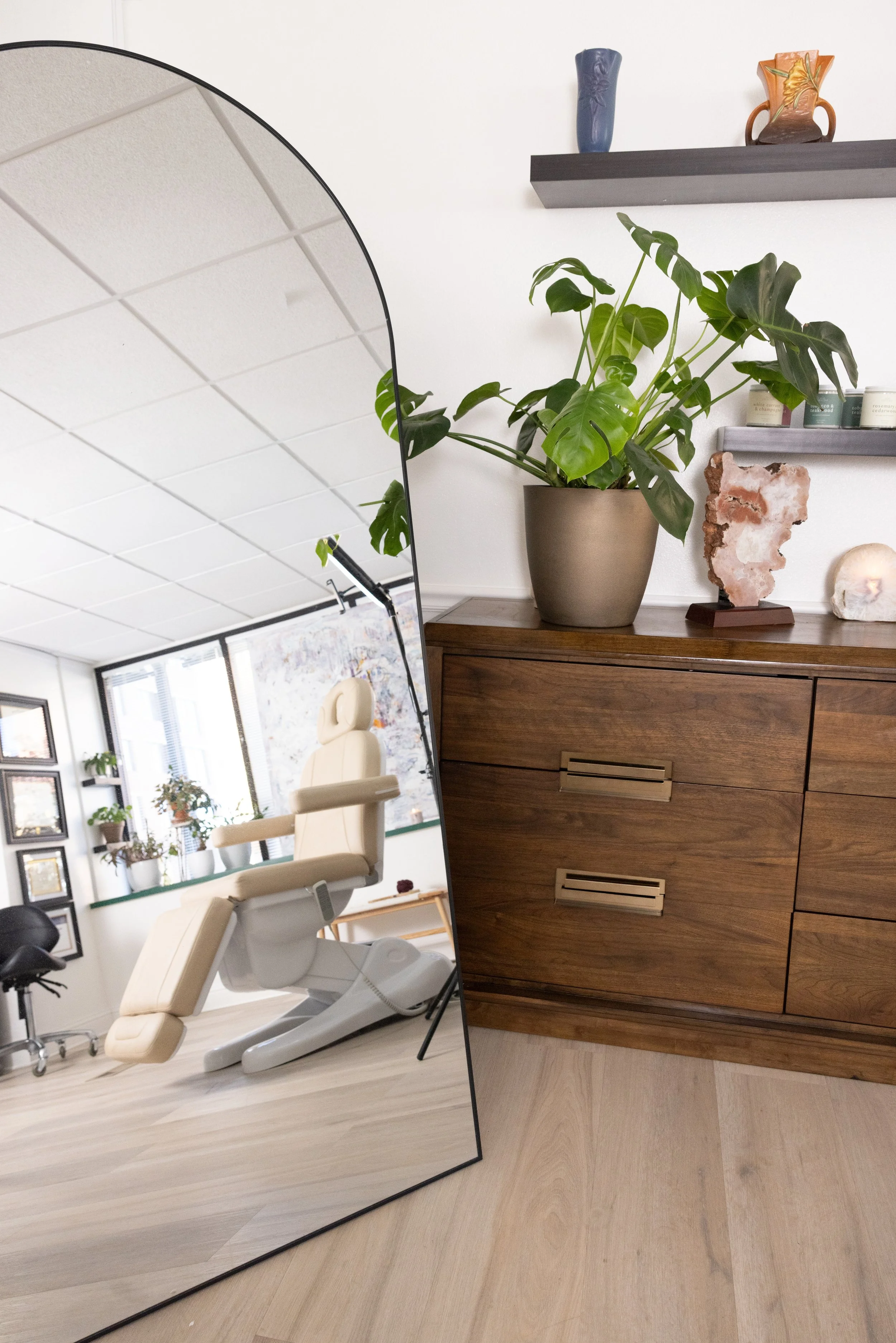 Large mirror reflecting Intuition Medical Aesthetics treatment. Next to the mirror is a wooden sideboard with a potted plant, geode, shell, and candles on floating shelves above.