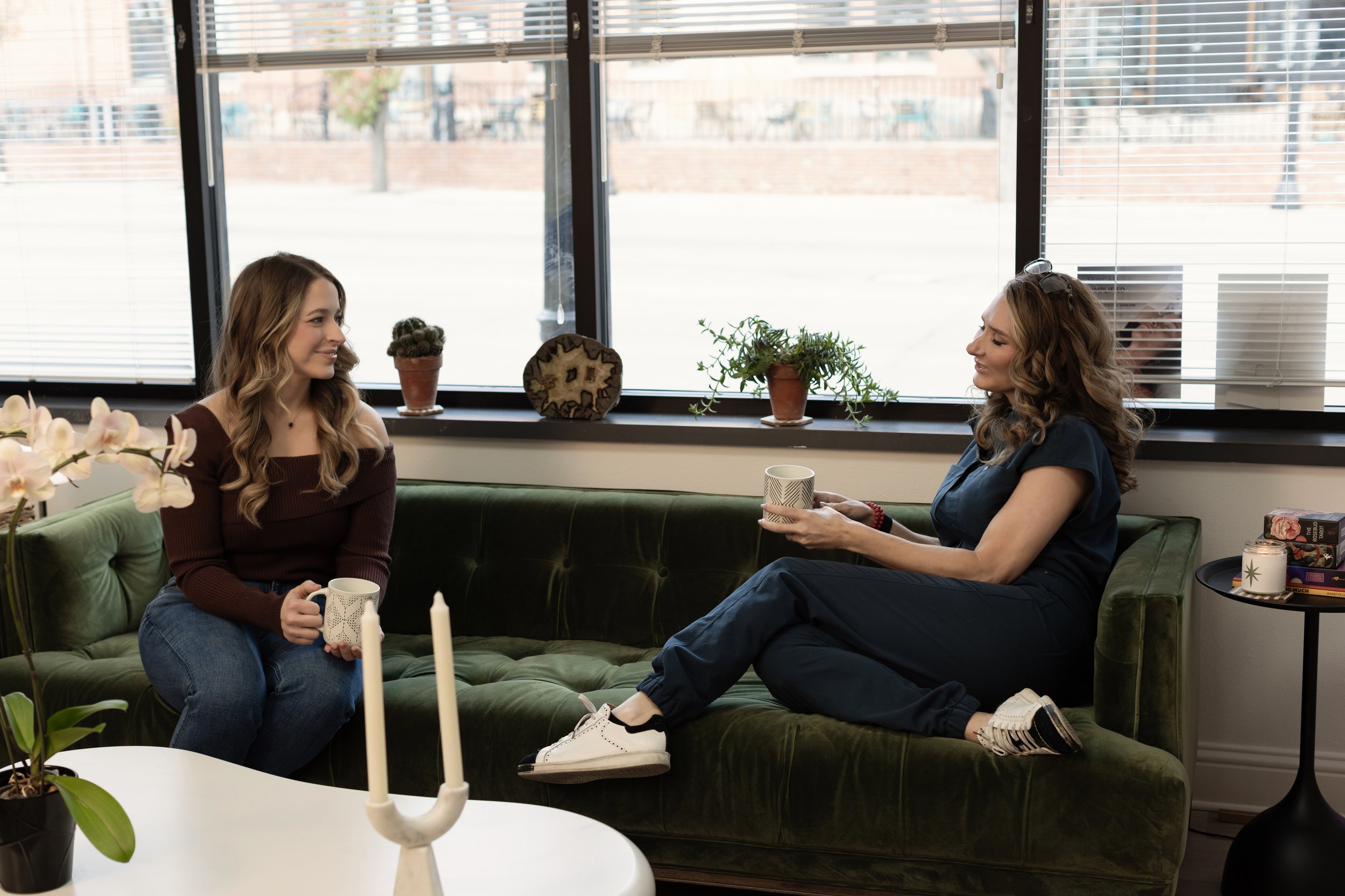 Two women are sitting on a green sofa in a sunlit room, having a conversation and sharing a coffee. One woman, with long wavy hair, is sitting cross-legged on the sofa, while the other, with shorter wavy hair, is sitting with her legs stretched out. They are surrounded by plants, candles, and decorative items on the windowsill and side table.