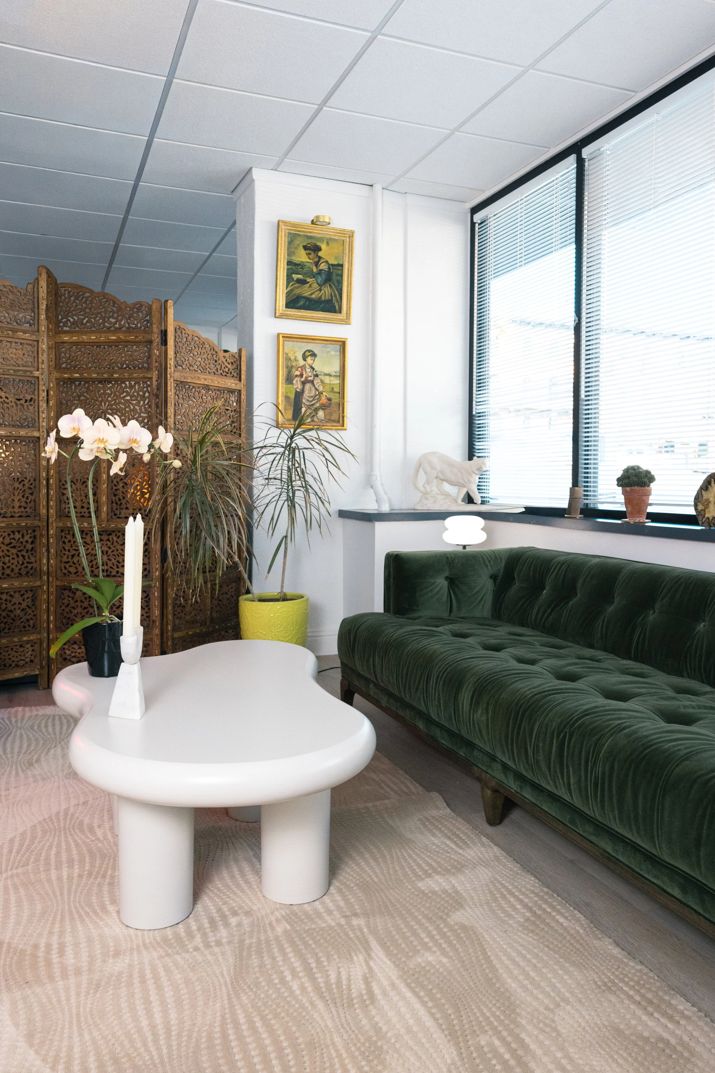 Living room with a green velvet sofa, white sculpted coffee table, potted plants in yellow and black planters, three framed paintings on the wall, a wooden folding room divider, and a window with blinds.