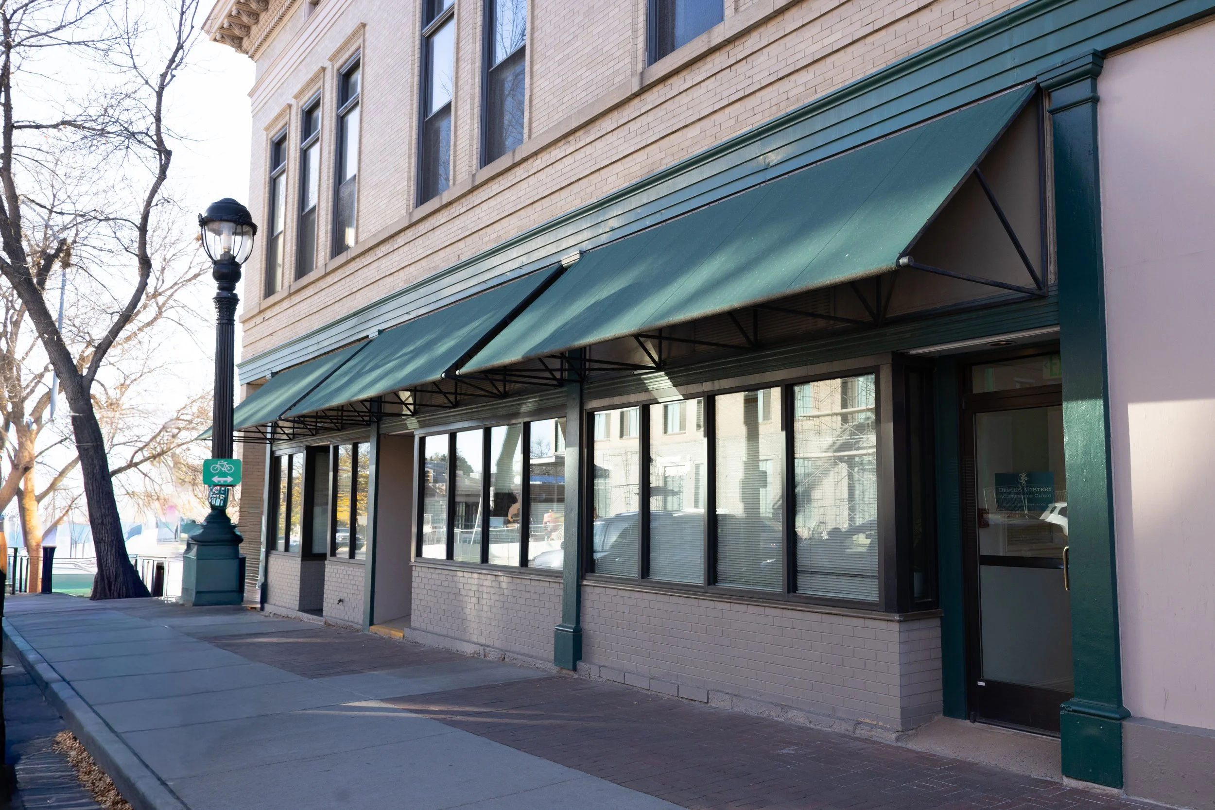 Storefront with green awnings, large glass windows, and a glass door. A park bench and a lamppost with a bike sign are visible on the sidewalk.