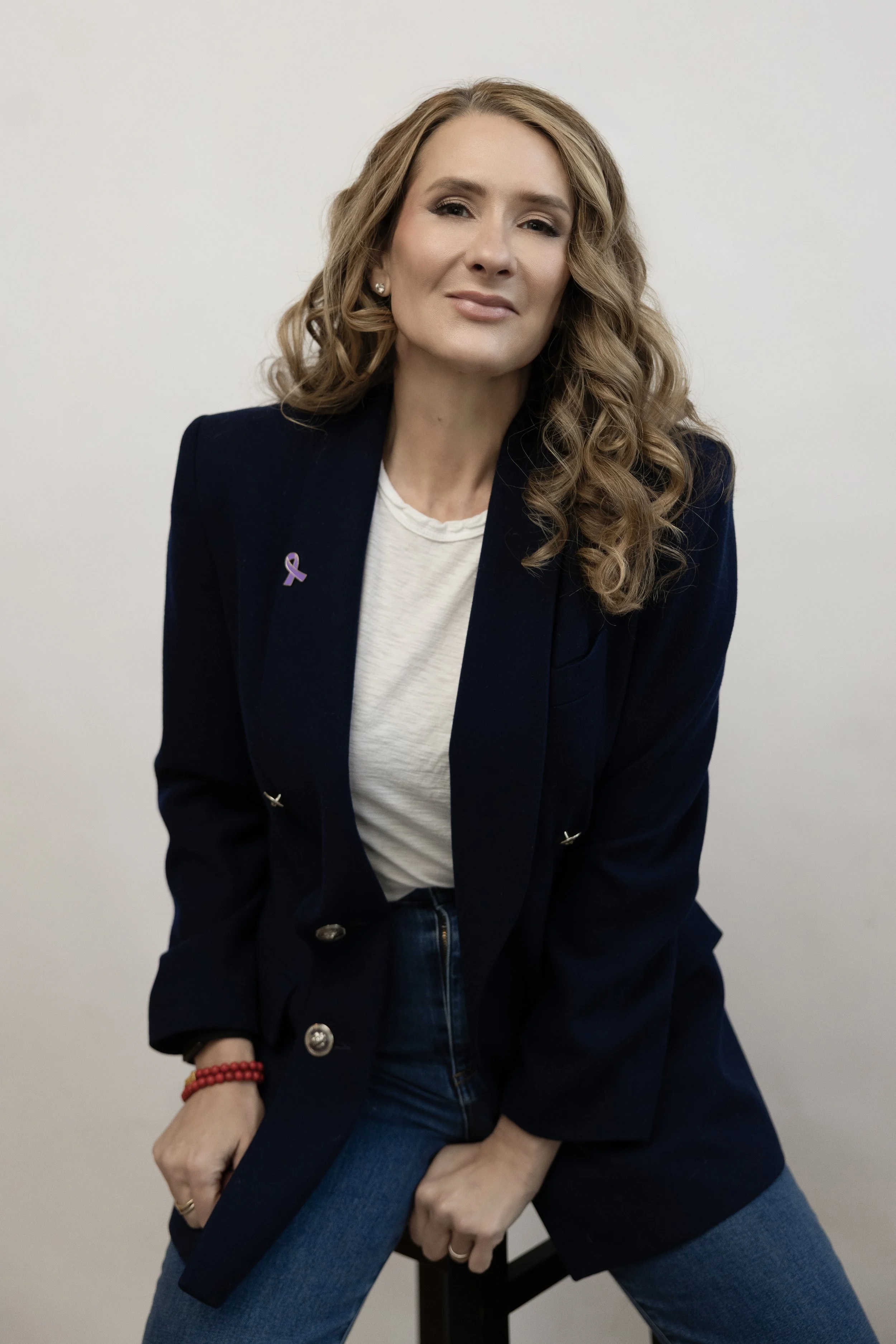 Woman with curly hair wearing a dark blazer and white shirt in front of a white wall.