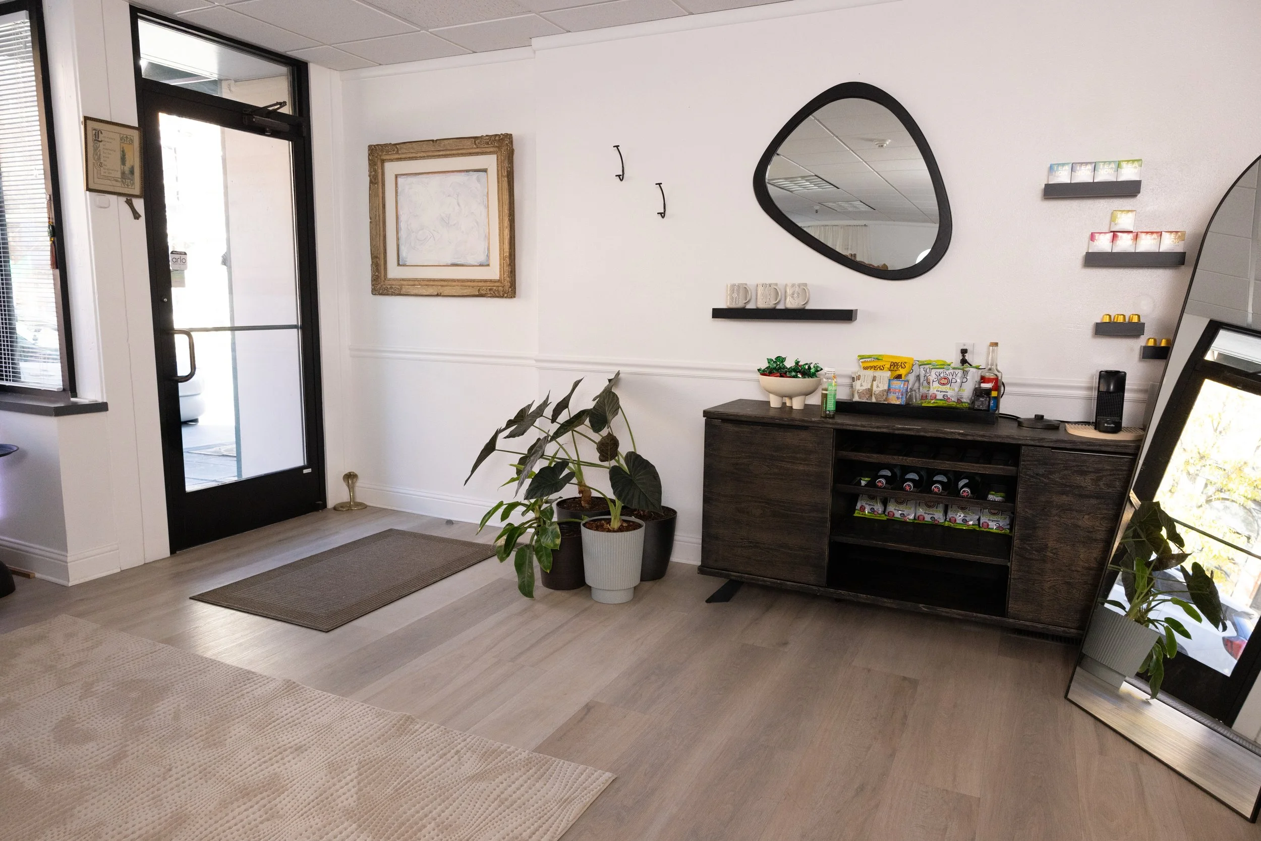 Lobby area with black door, framed artwork, potted plants, a dark wood sideboard with snack items and drinks, a round mirror, and shelves with small items, all on light wooden flooring.