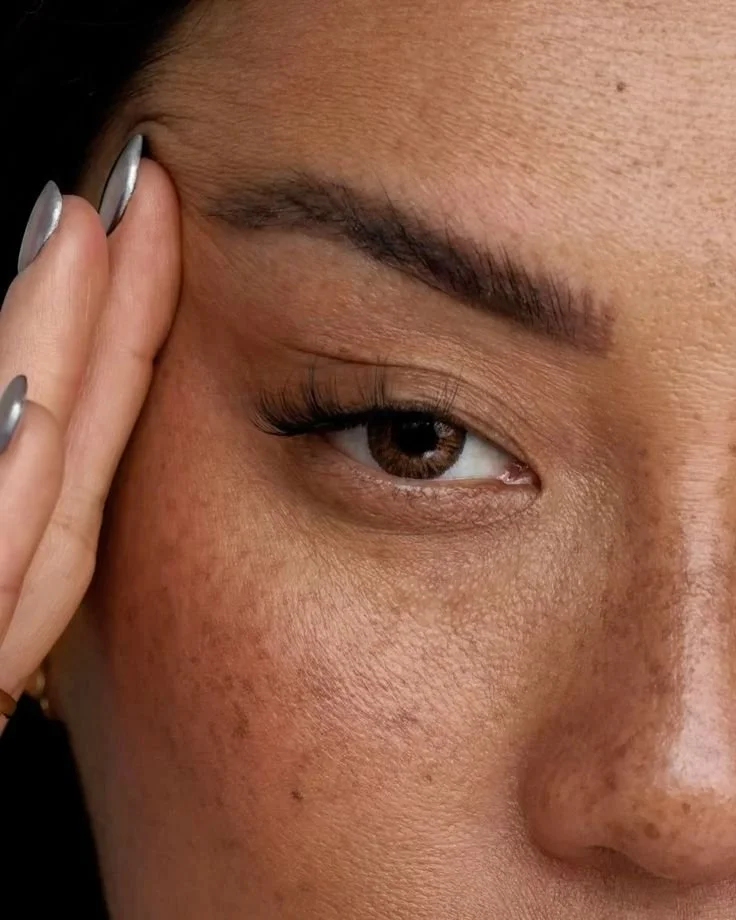 Close-up of a woman's face focusing on her eye, eyebrow, and skin texture, with her hand gently touching her forehead.