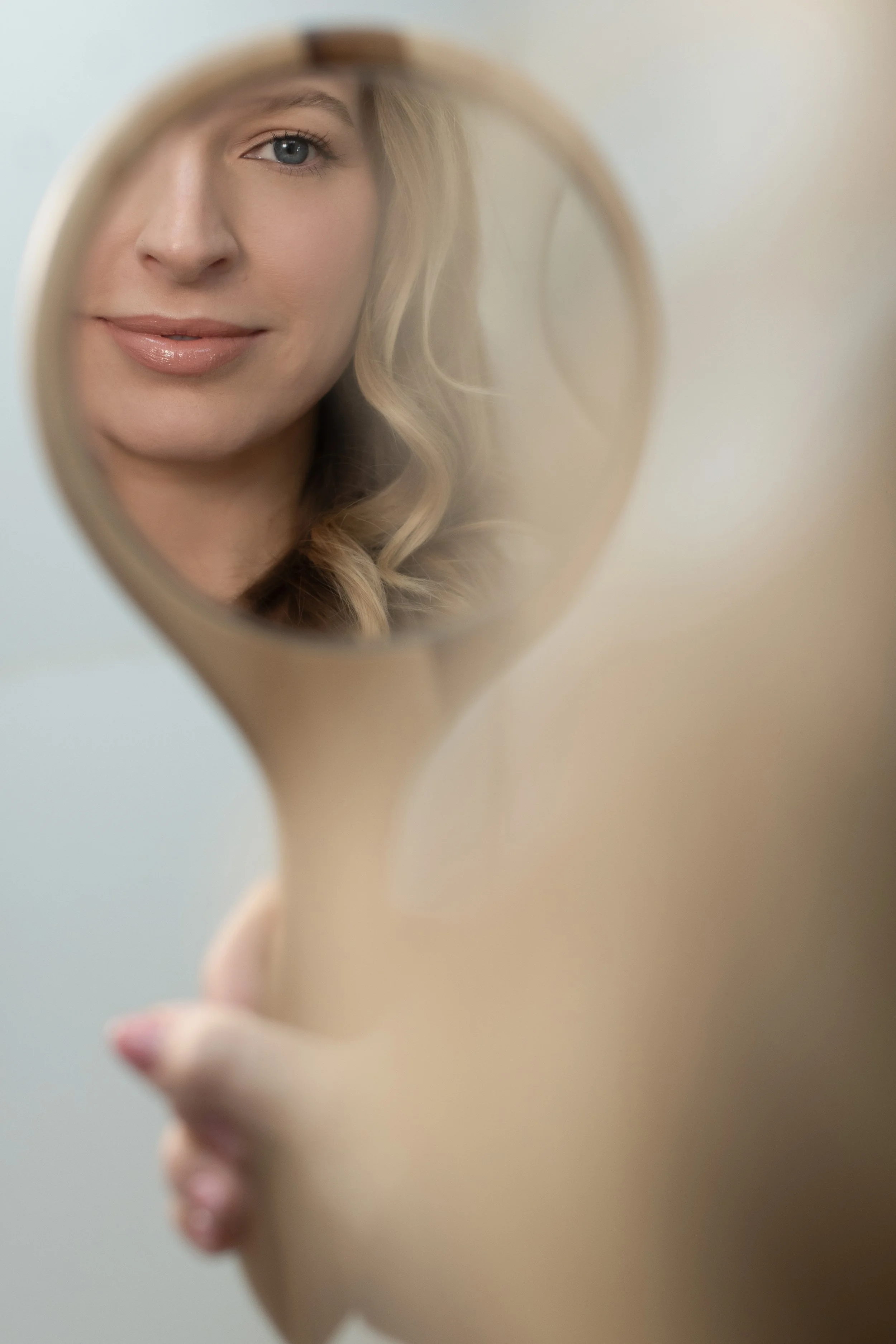 Close-up of a woman looking into a hand mirror, smiling with blonde curly hair, light skin, and wearing natural makeup.