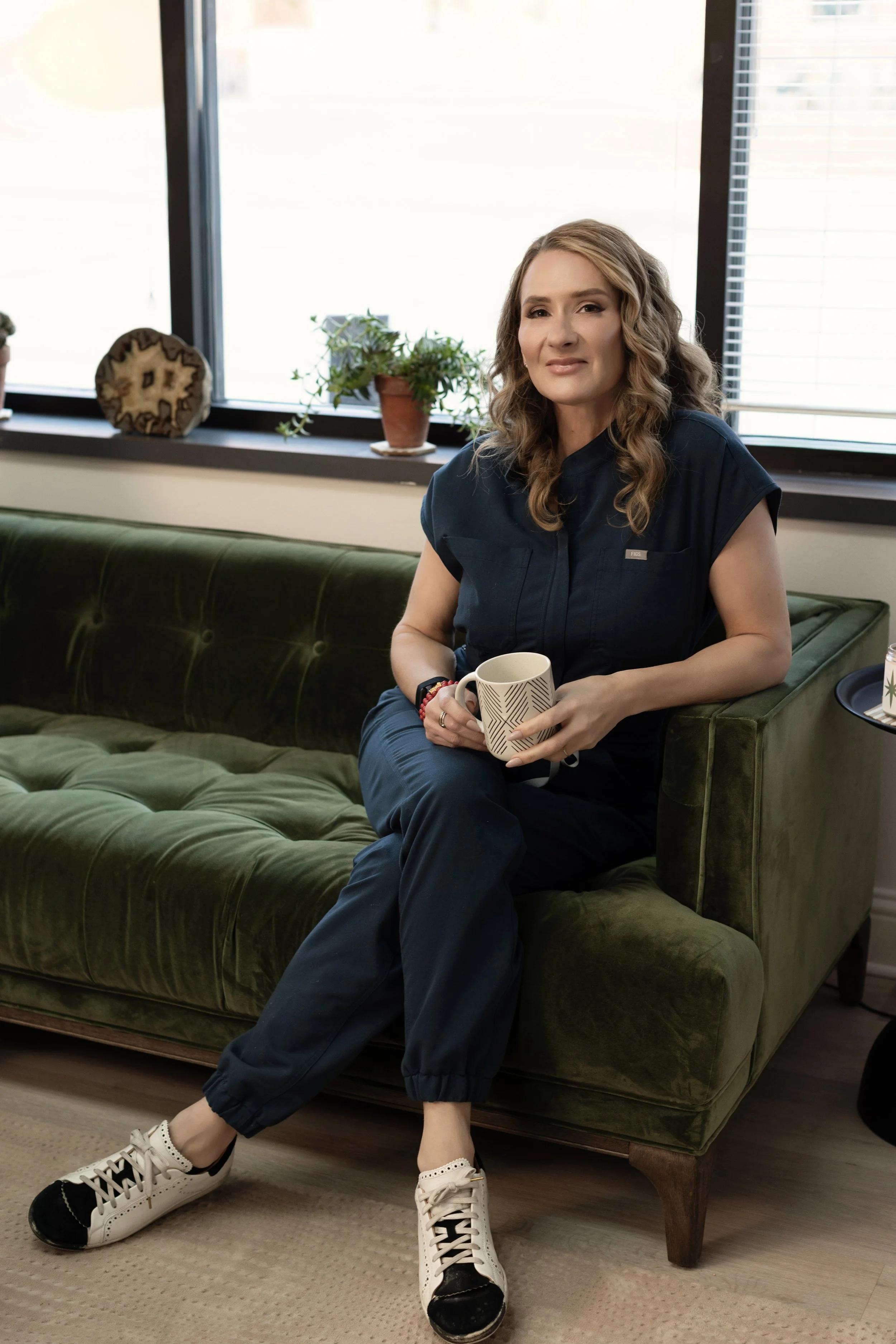 Injector Nissa, wearing navy scrubs, sitting on a green velvet sofa and holding a mug at Intuition Medical Aesthetics
