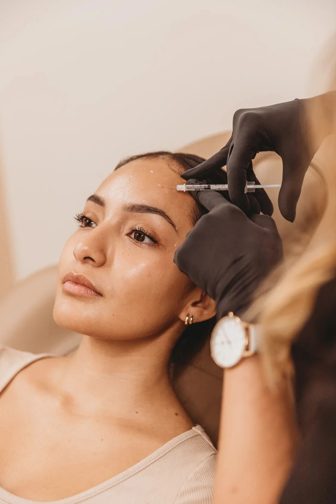 A woman with light skin and brown hair is receiving an injection in her forehead from a professional wearing black gloves.