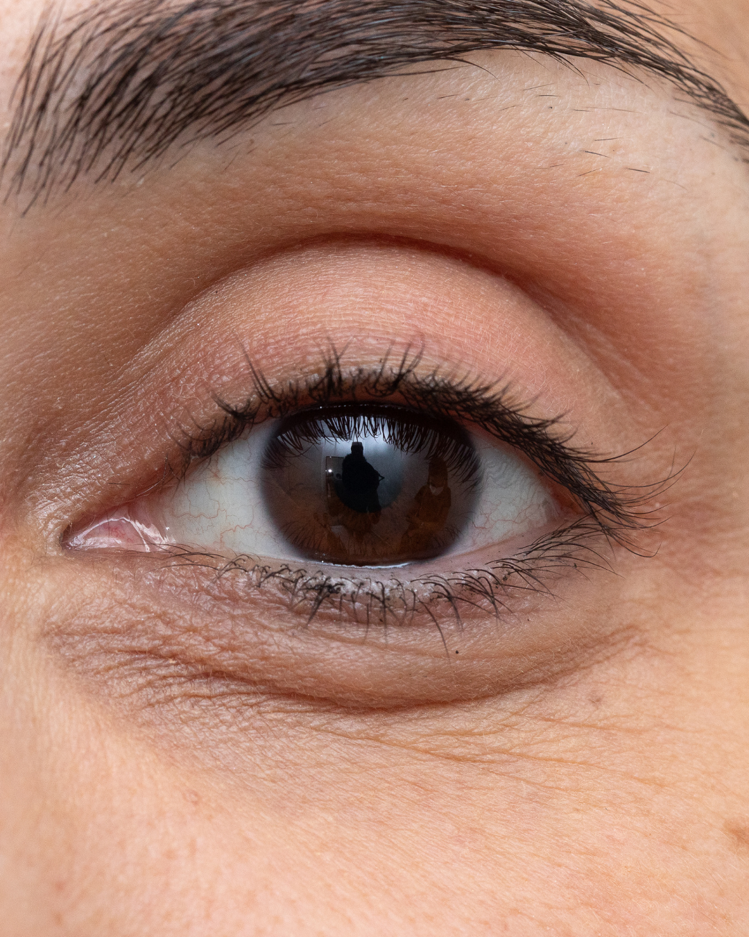 Close-up of a human eye with brown iris and long eyelashes, showing reflection of a person taking a photo with a camera.