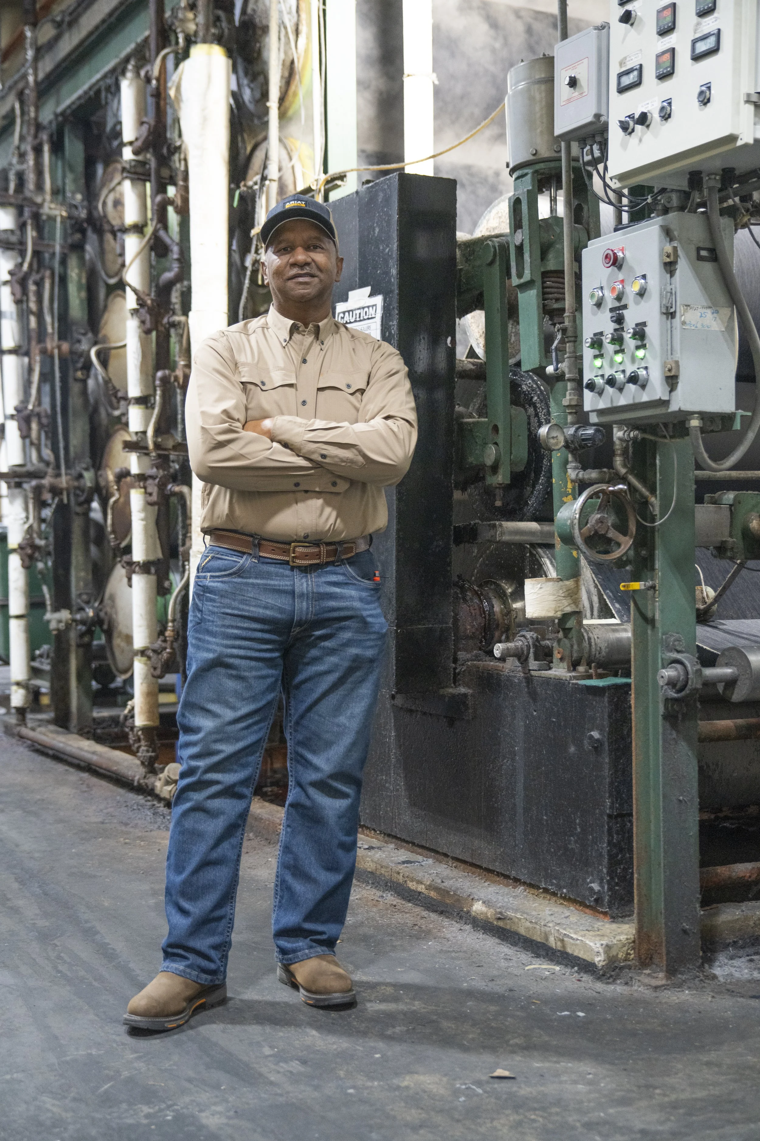 A man in jeans and a tan work shirt with arms crossed, standing in front of industrial machinery in a factory setting.