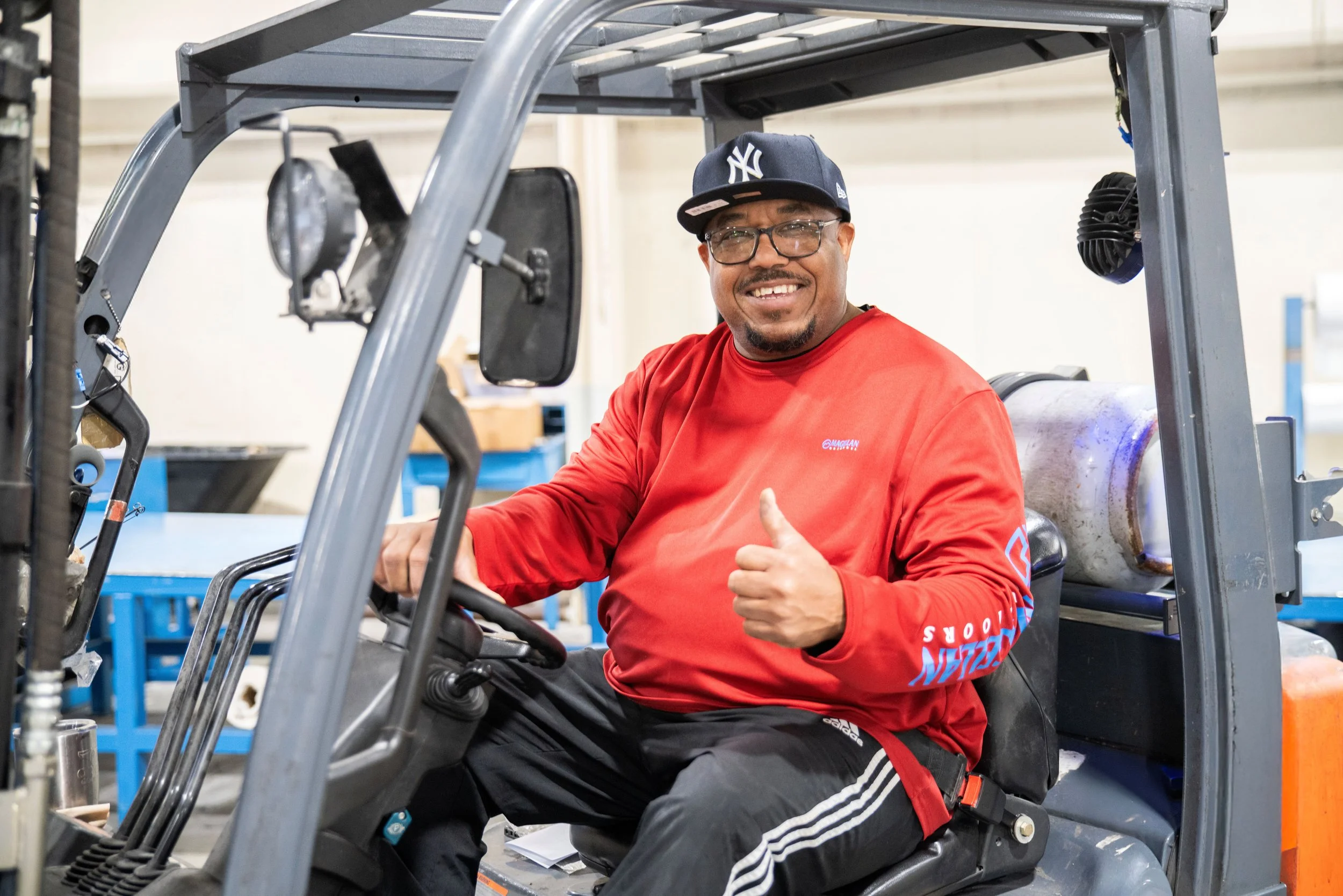 A man wearing glasses, a New York Yankees cap, and a red long sleeve shirt sitting in a golf cart, giving a thumbs-up and smiling.