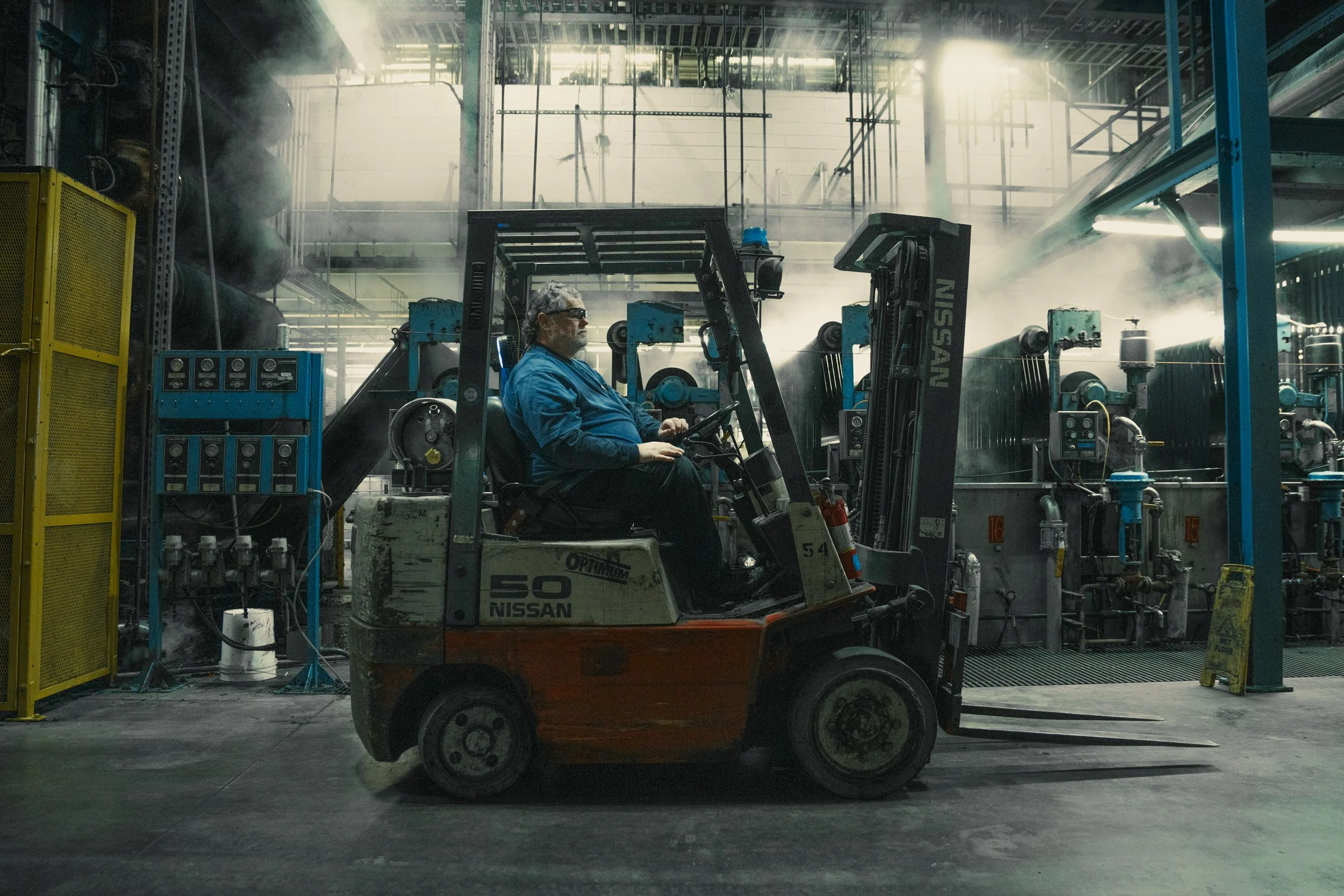 A man operating a forklift inside an industrial warehouse filled with machinery and piping, with sunlight streaming through windows.