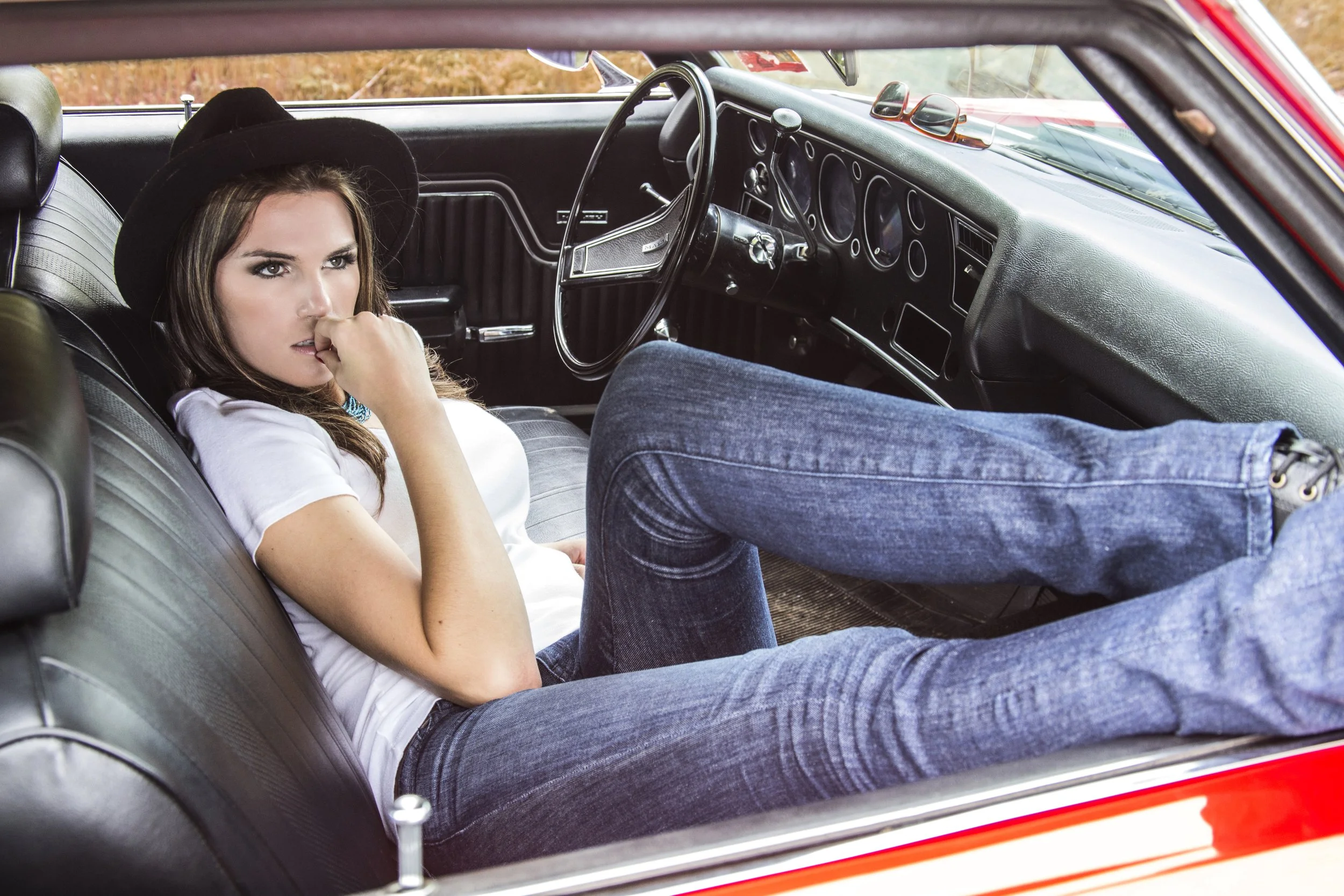 Woman in a black hat sitting in the backseat of a vintage red car, with a man lying across her lap, in an outdoor setting.