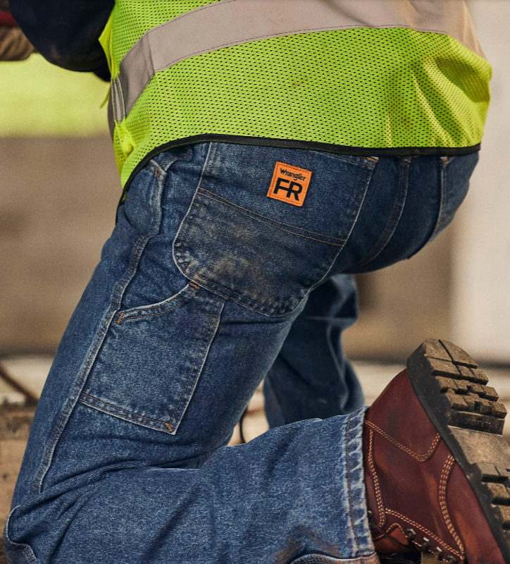 Close-up of a construction worker wearing jeans with a Wrangler FR patch, high-visibility safety vest, and brown work boots.