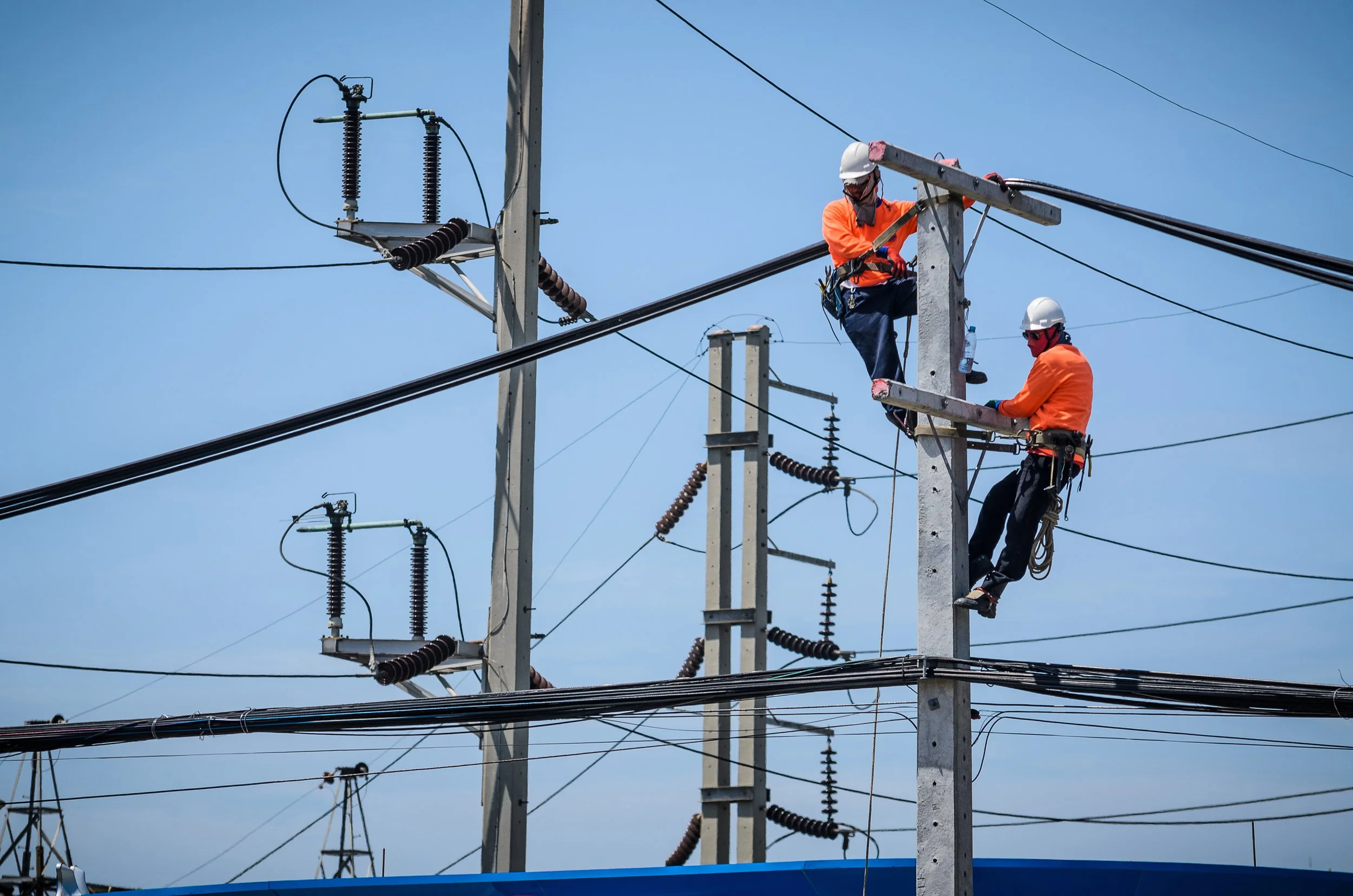 Two utility workers wearing safety helmets and orange shirts working on electrical power lines on tall utility poles, with a clear blue sky in the background.