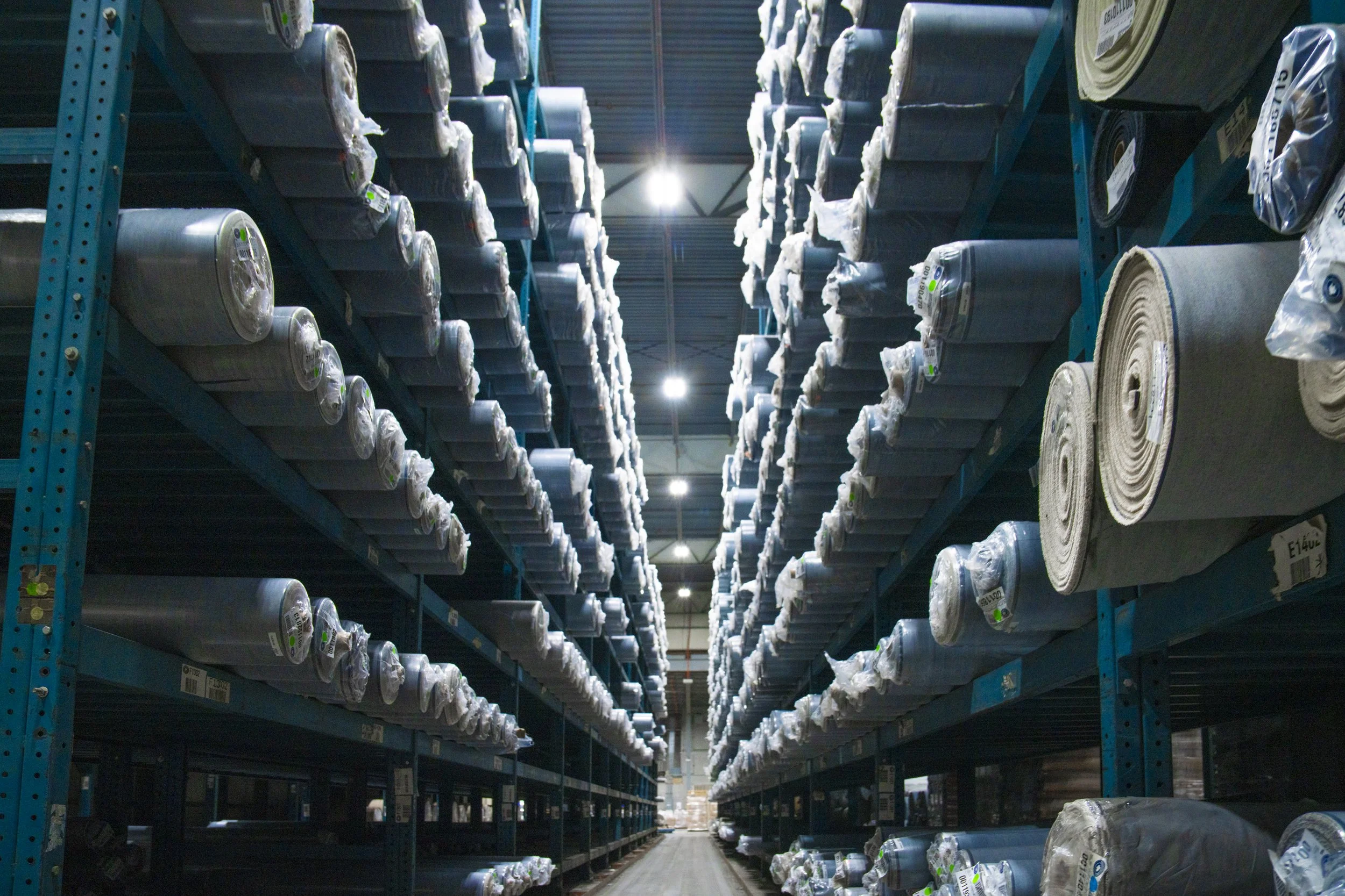 View of tall blue metal shelves filled with large rolls of fabric stored vertically in a warehouse, with bright overhead lighting.
