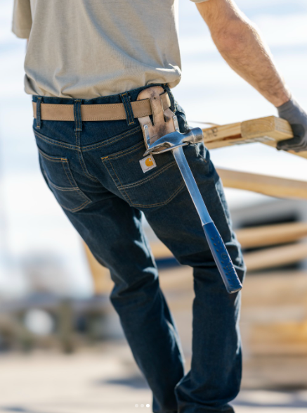 Close-up of a person holding a wooden plank and a hammer, wearing jeans and a tool belt on a construction site.