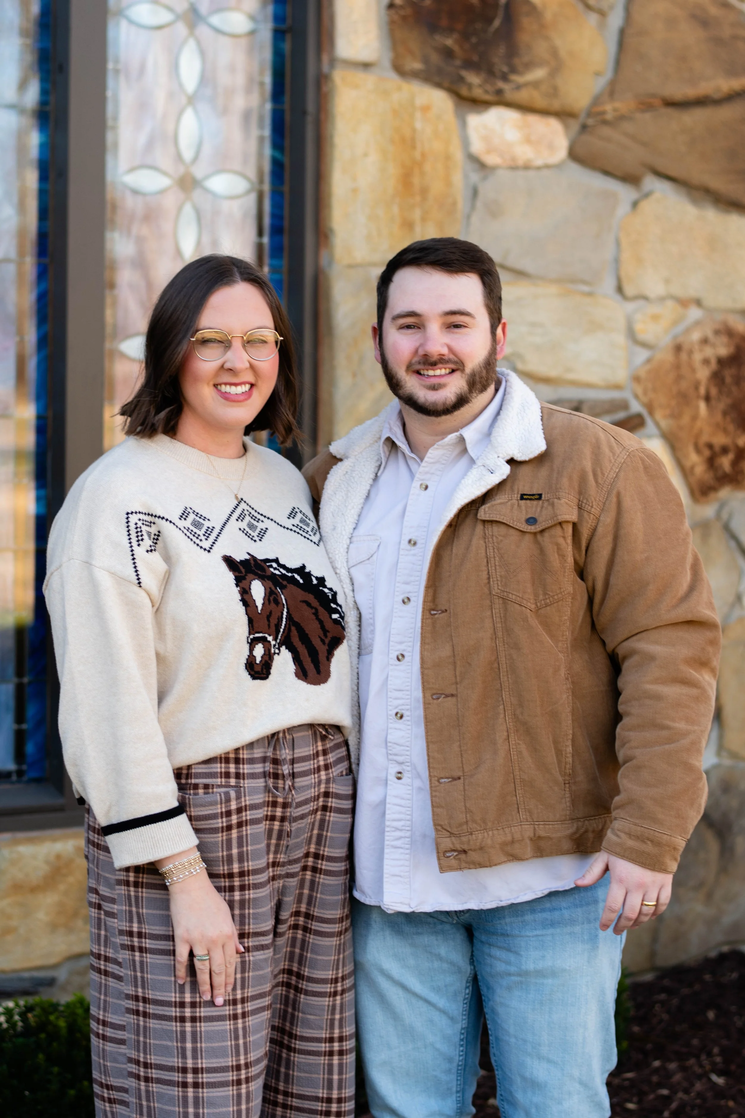 A smiling couple standing outside in front of a stone wall. The woman has shoulder-length brown hair, glasses, and is wearing a cream sweater with a horse design, plaid pants, and jewelry. The man has short dark hair and a beard, and is wearing a brown jacket over a white shirt and light jeans.