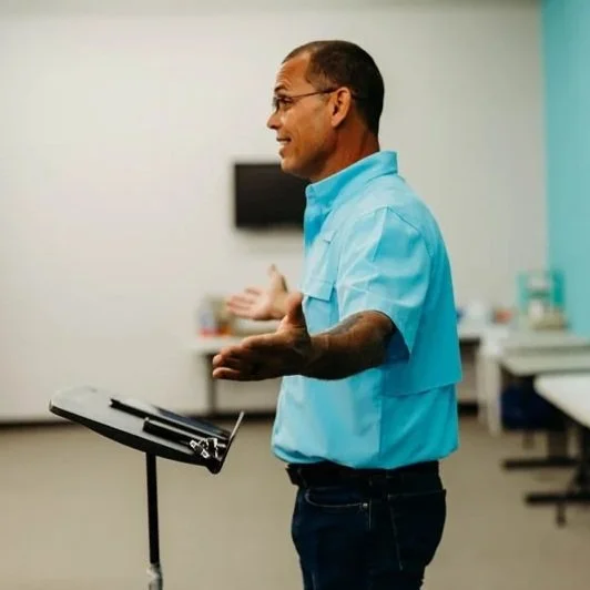 A man dressed in a blue shirt, standing in a medical or clinical setting, speaking and gesturing with his hands, with medical equipment and beds in the background.