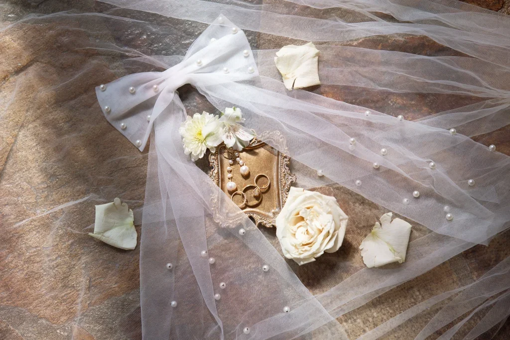 Wedding rings on an ornate silver tray surrounded by white roses, petals, pearl embellishments, and tulle fabric.