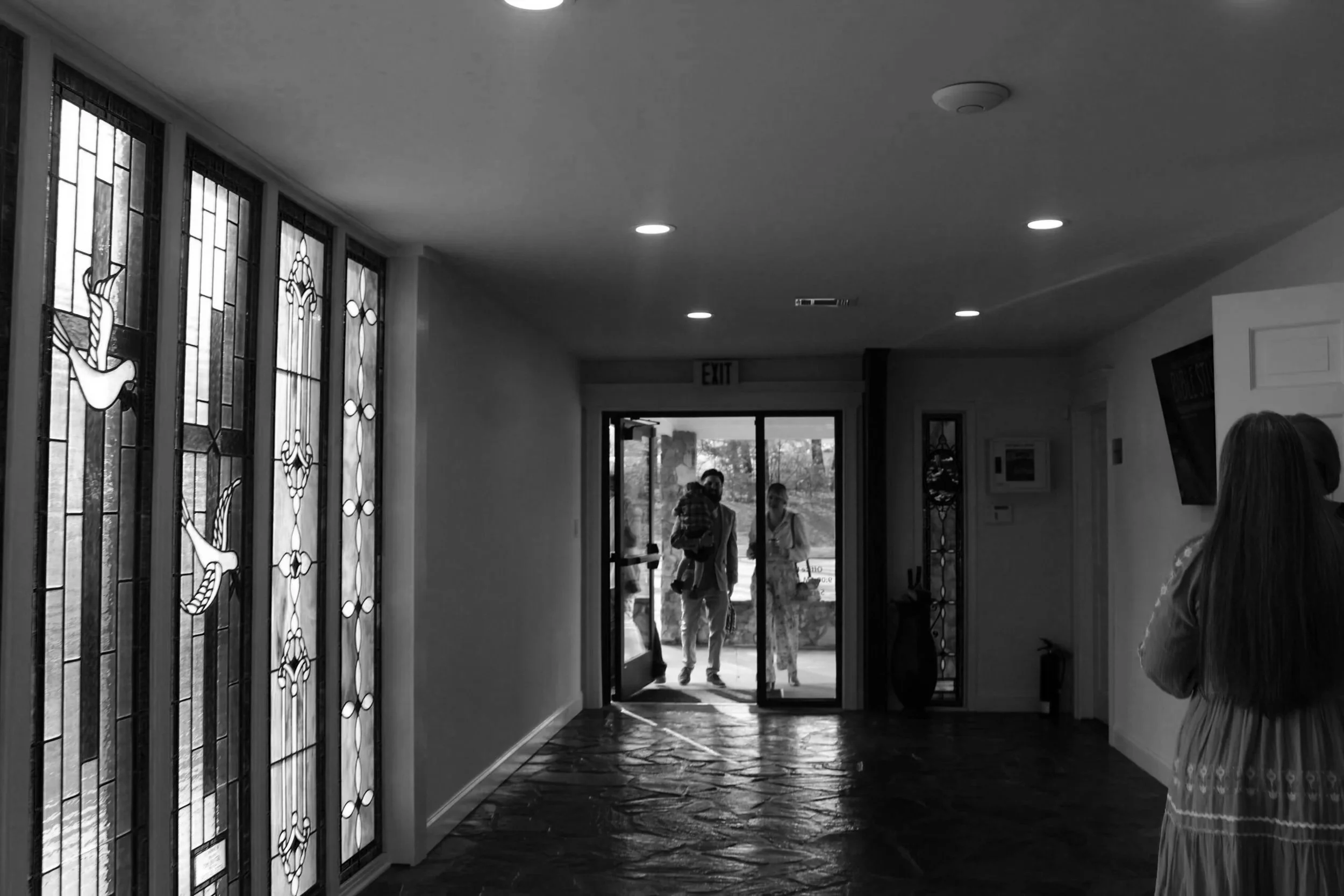 People entering a building through a glass door, seen from inside near stained glass windows.