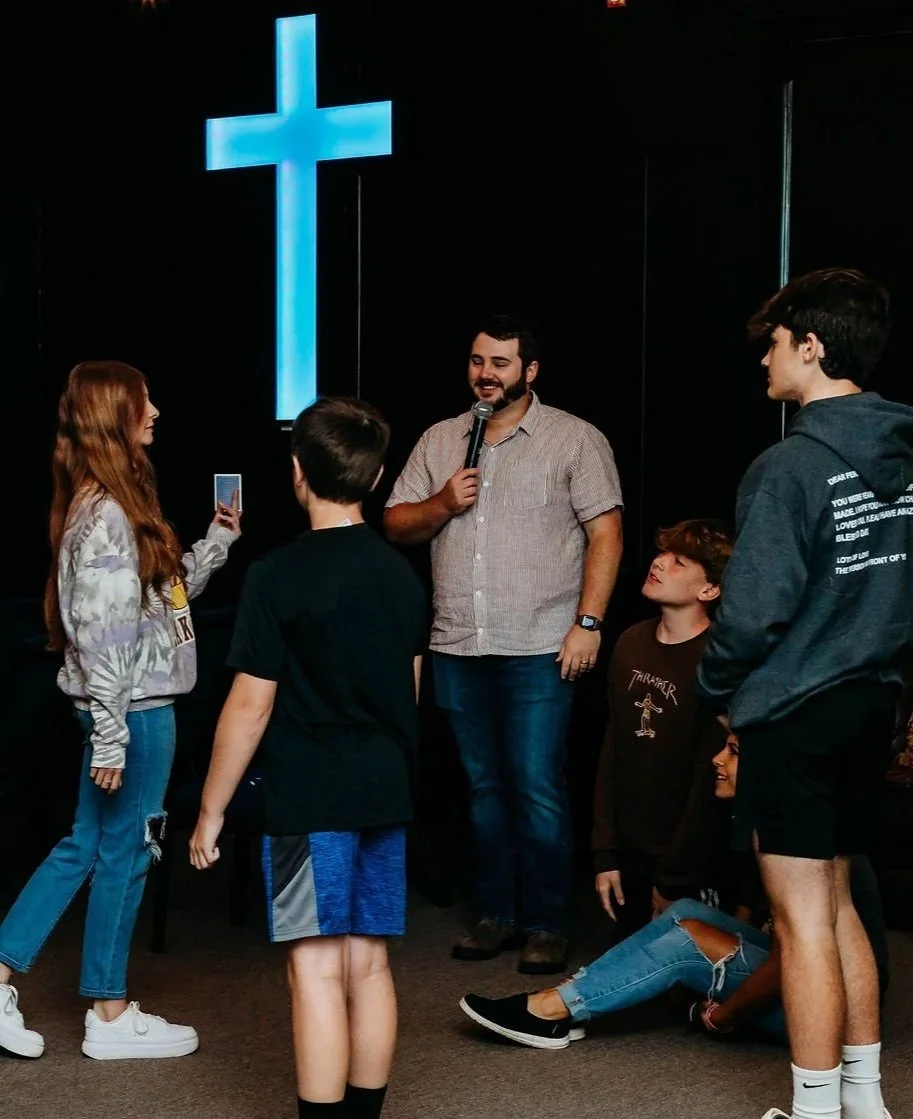 Group of teenagers and a man with a microphone gathered around a woman taking a photo, in front of a large illuminated Christian cross.