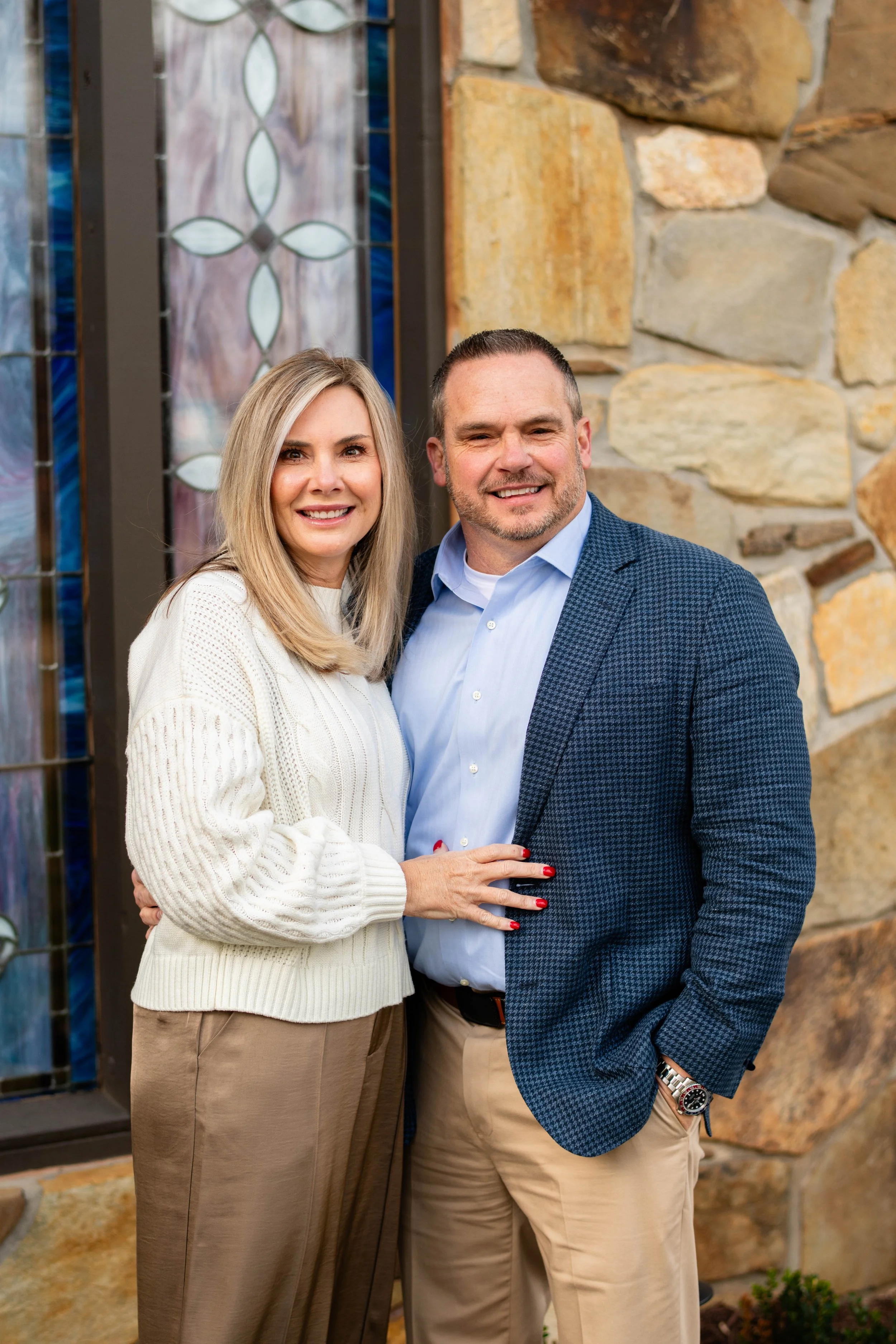 A smiling middle-aged couple standing close together in front of a stone wall with stained glass windows, the woman has blonde hair, light-colored sweater, and beige pants, and the man has short hair, a beard, a blue patterned blazer, and khaki pants.