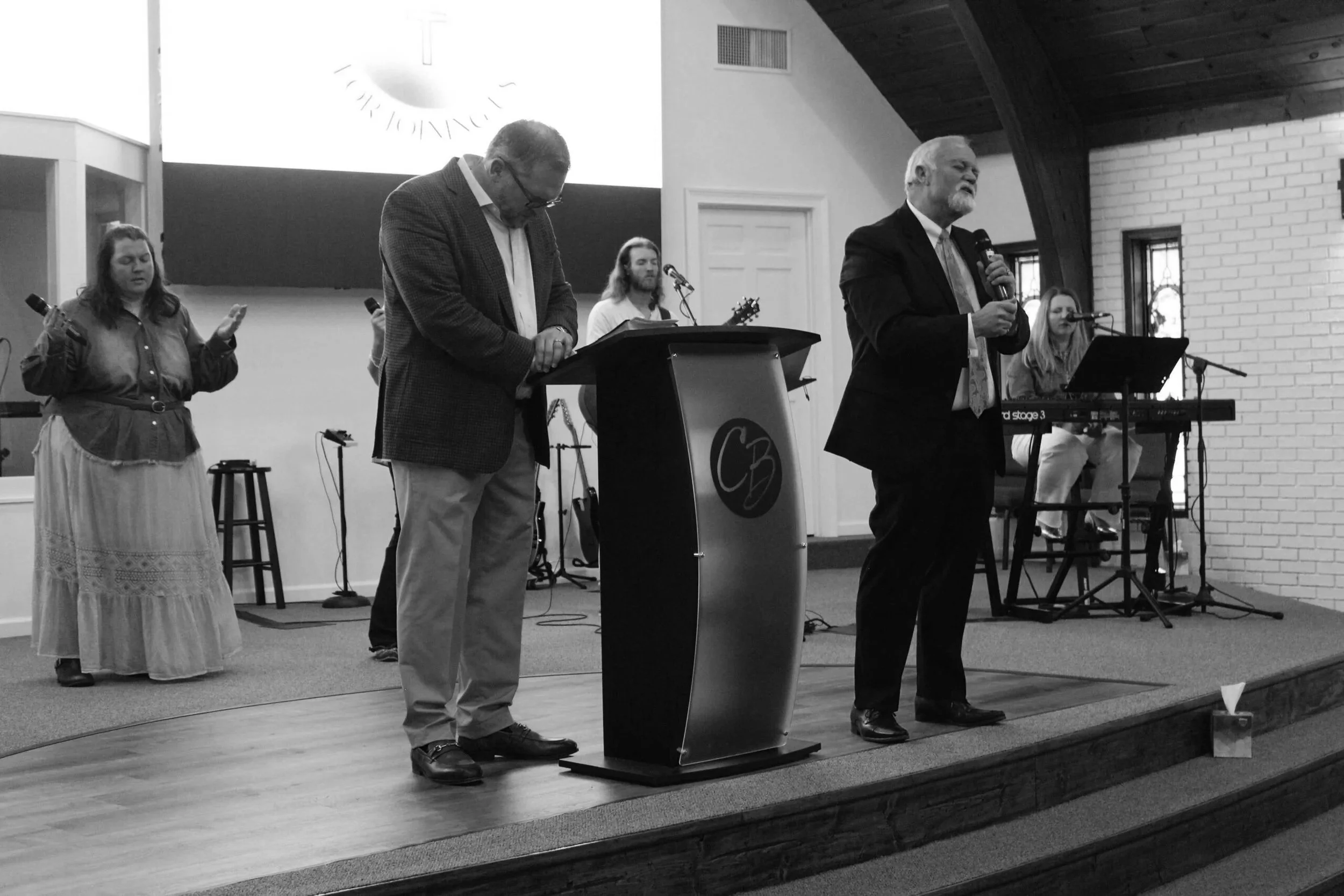A group of people on stage during a church service or event. One man is speaking into a microphone, while another is kneeling at a podium with a logo. A woman with a microphone and a long skirt, and two musicians with guitars and a keyboard are also present.