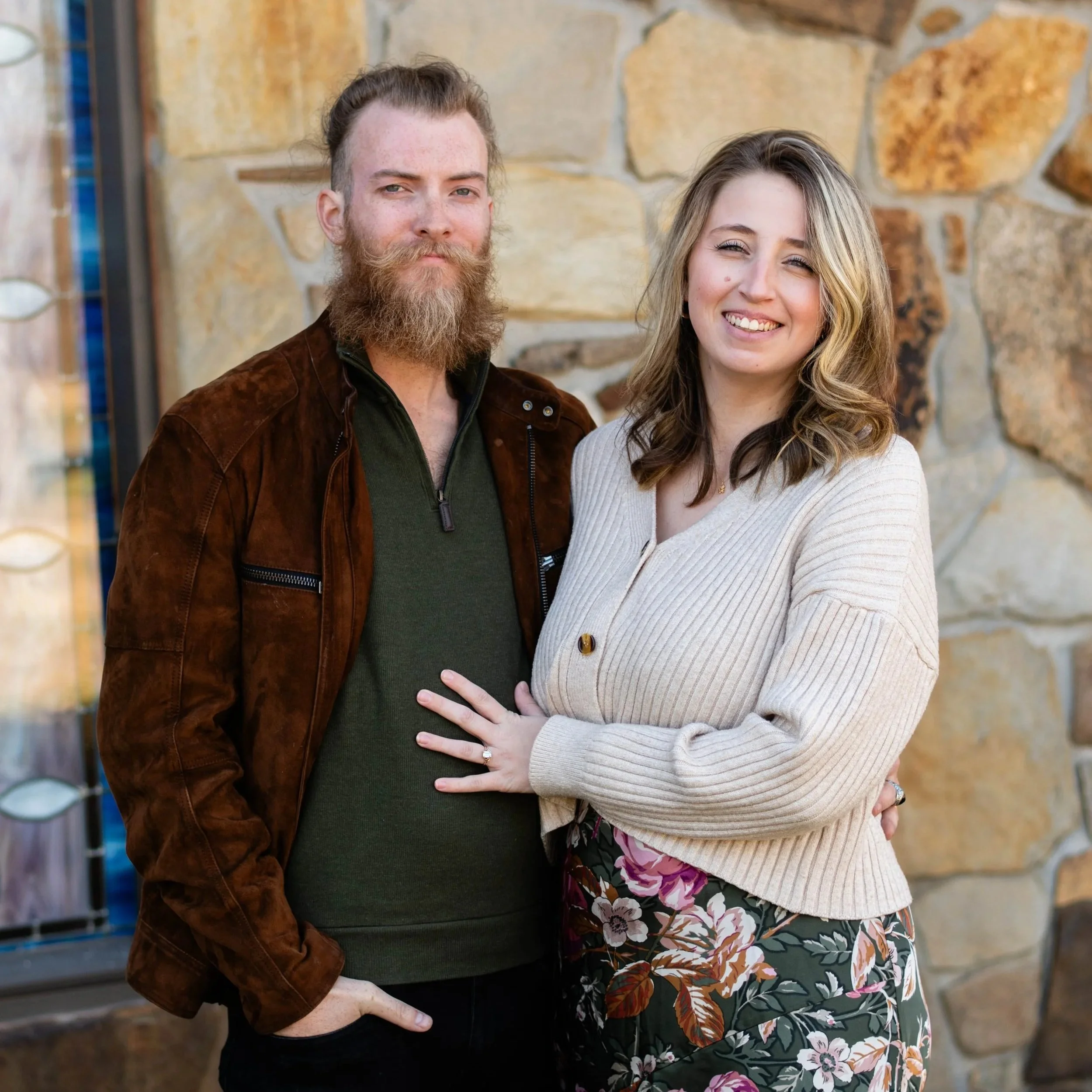 A man with a beard and a woman with shoulder-length hair smiling and posing together in front of a stone wall.
