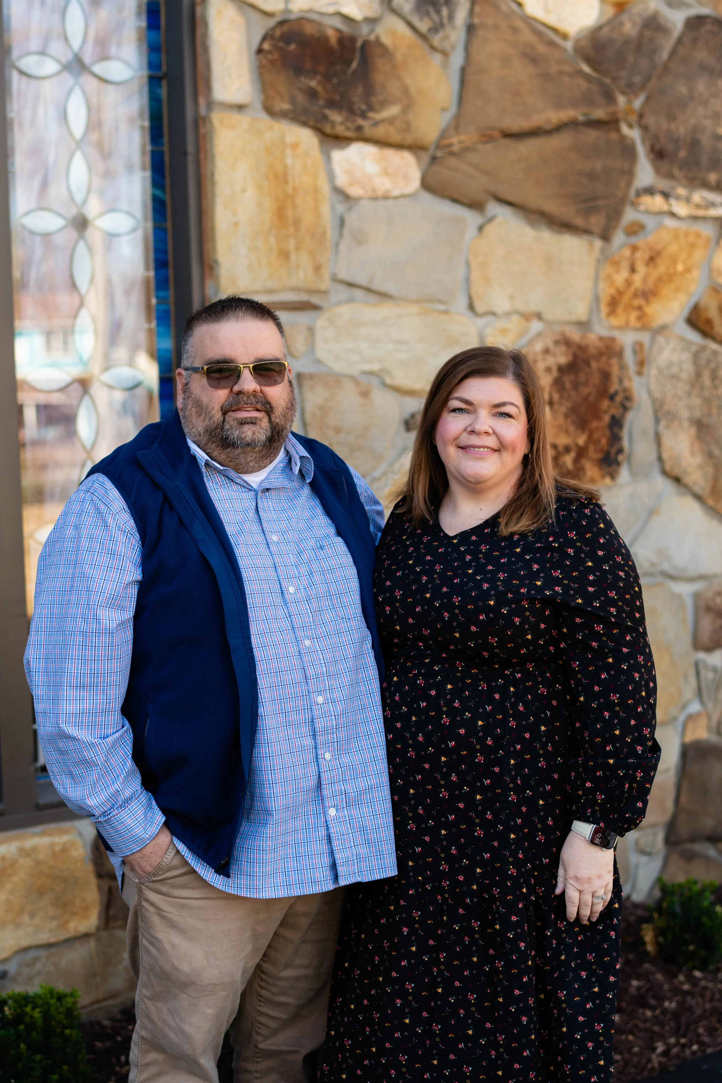 A man and woman standing outside in front of a stone wall, smiling at the camera.