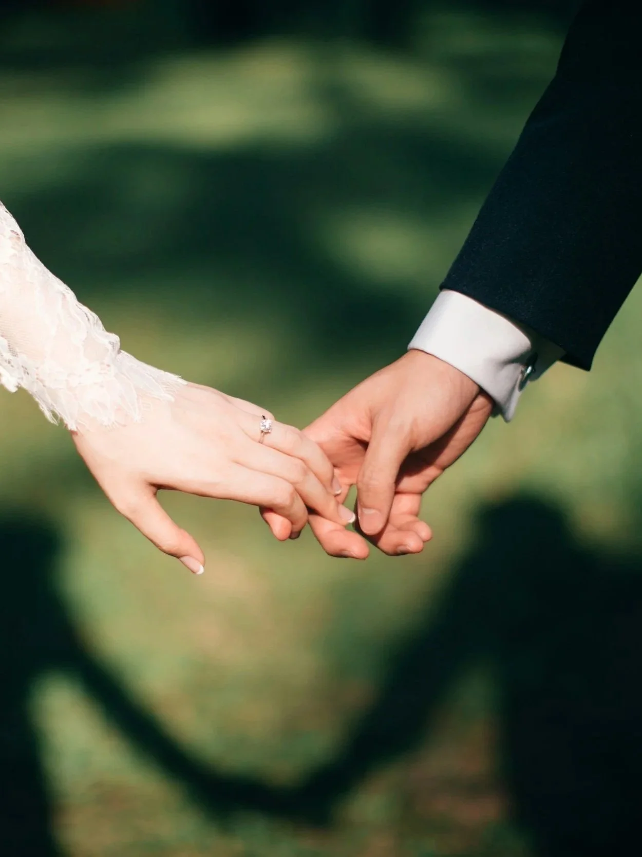 Close-up of a bride and groom holding hands, with the bride wearing a white lace dress and a wedding ring, and the groom wearing a dark suit with a white shirt, outdoors with a blurred green background.