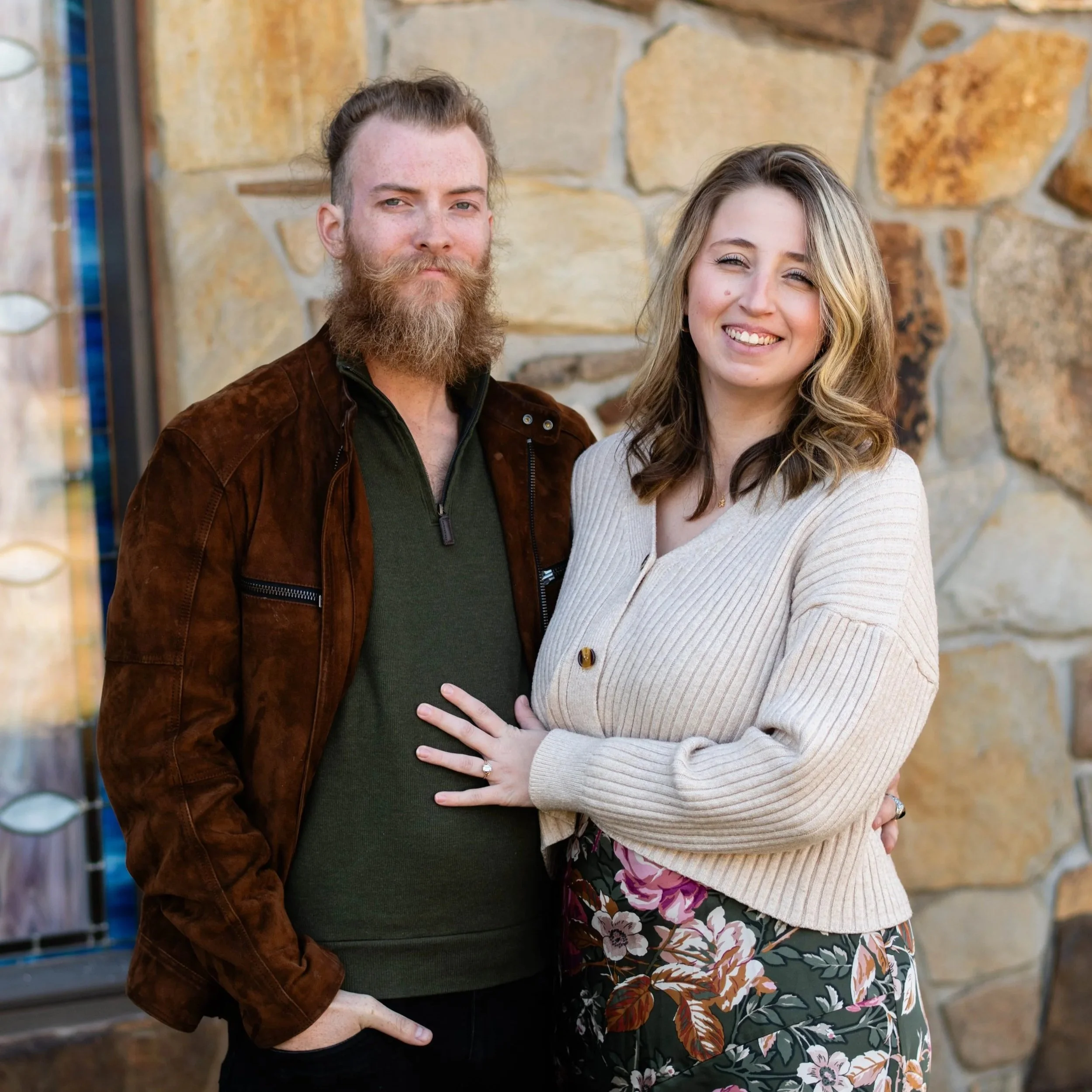 A man with a beard and a woman with wavy hair smiling and standing close together outside against a stone wall.
