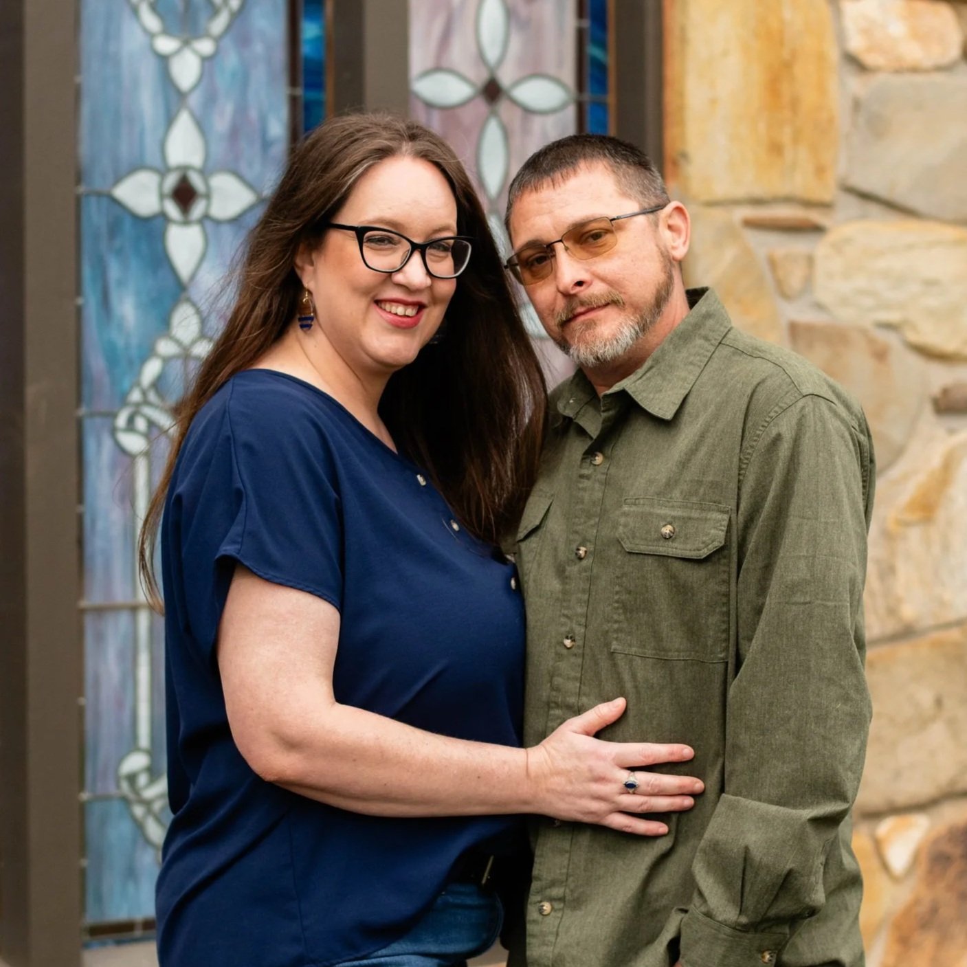 A smiling woman with long brown hair, wearing glasses and a royal blue shirt, standing close to a serious-looking man with short brown hair, beard, and glasses, wearing a green button-up shirt, in front of stained glass windows and a stone wall.