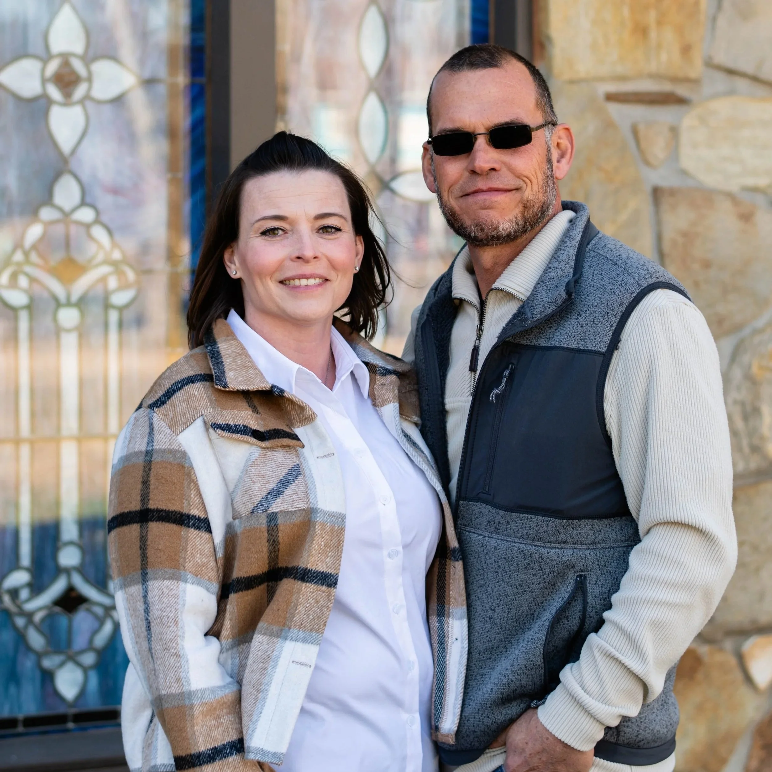 A man and woman standing outside, smiling at the camera, with a decorative window and stone wall in the background.