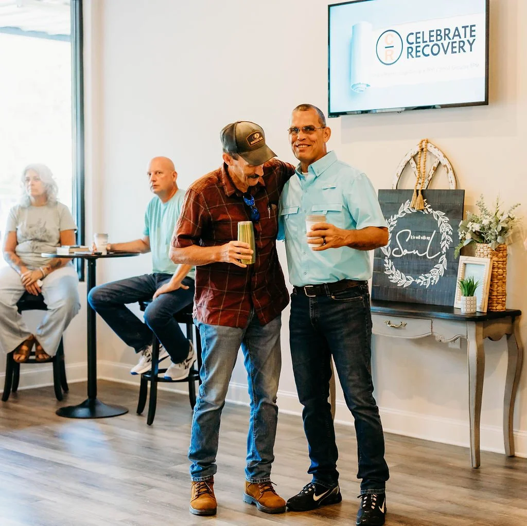 Two men standing close together, smiling and holding drinks at a gathering. In the background, three other people are sitting at a table. A large screen on the wall displays 'Celebrate Recovery,' and there are decorative elements on a table nearby, including a sign that says 'Psalm' with a wreath of leaves.