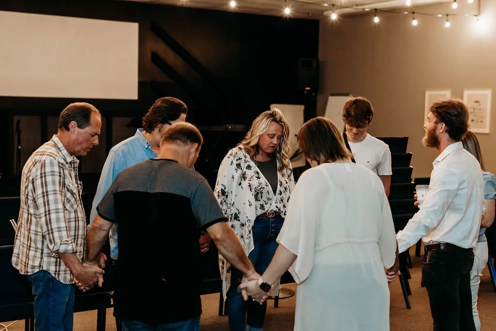 Group of people holding hands in prayer during a gathering in a dimly lit room.